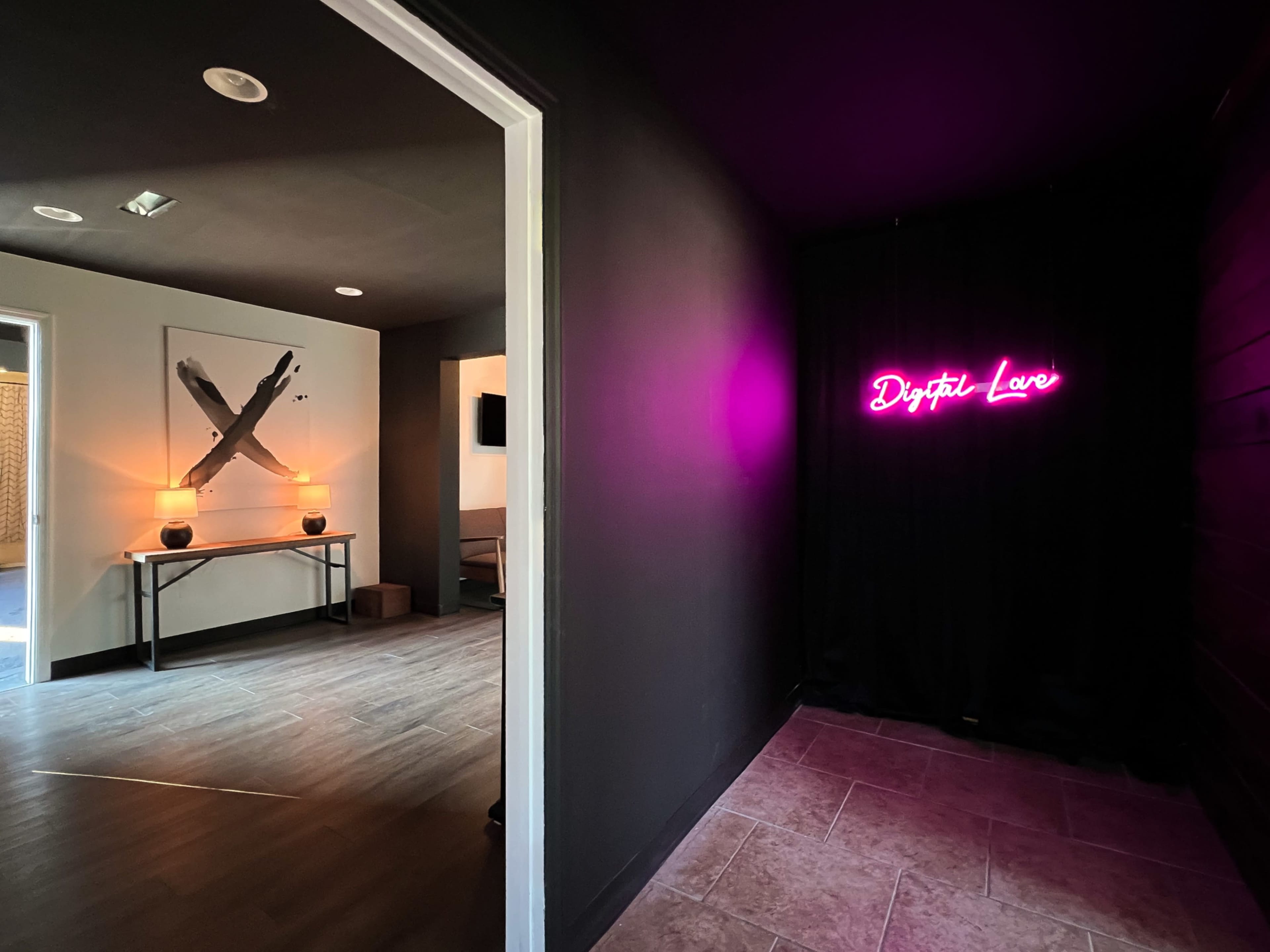 A dimly lit hallway leading to a black curtain with a neon sign that reads "Digital Love," while a room with decor and a table is visible through an open door to the left.
