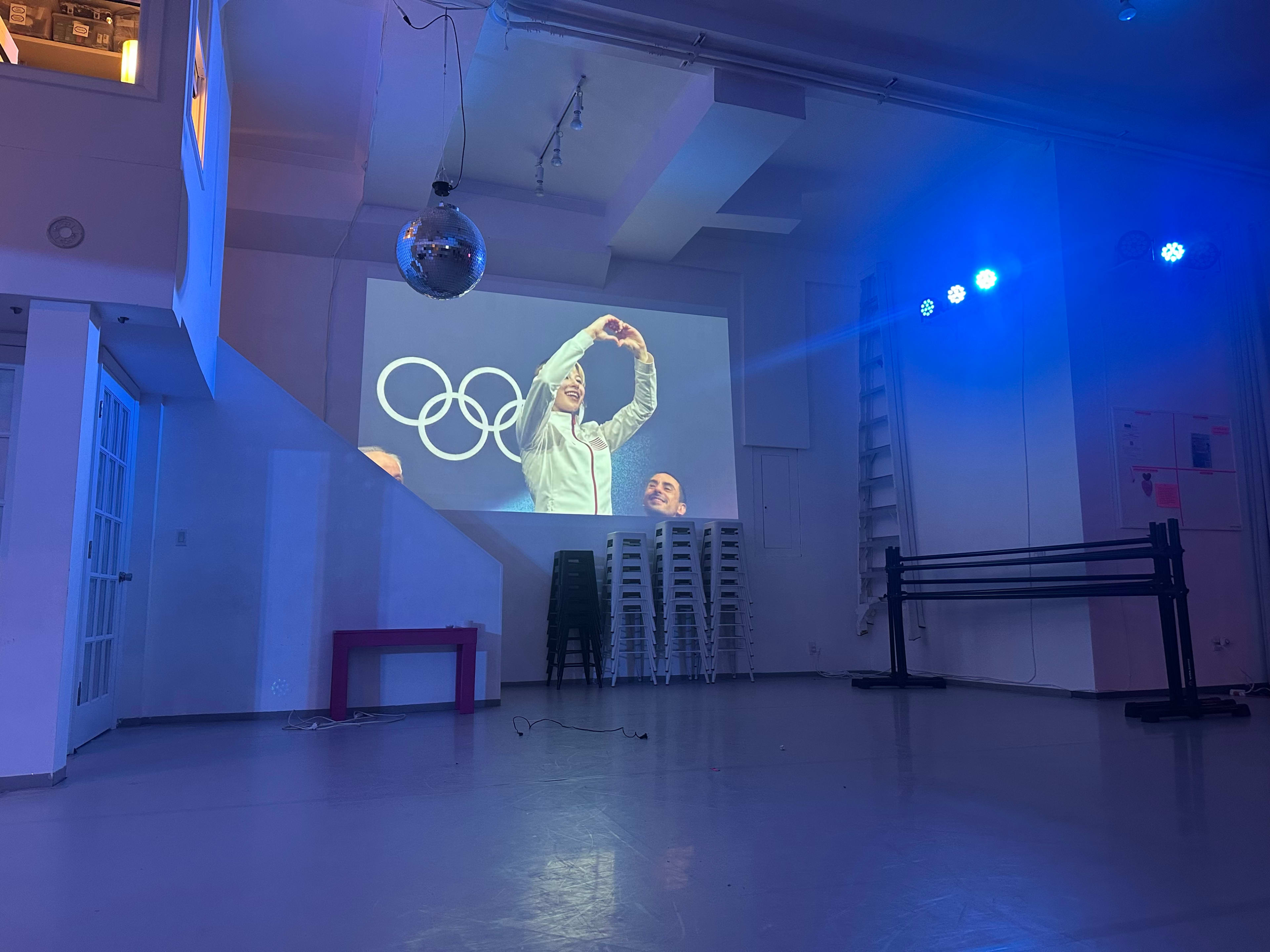 A room with a large projection of a person celebrating at the Olympics is illuminated by blue lighting and features a disco ball.