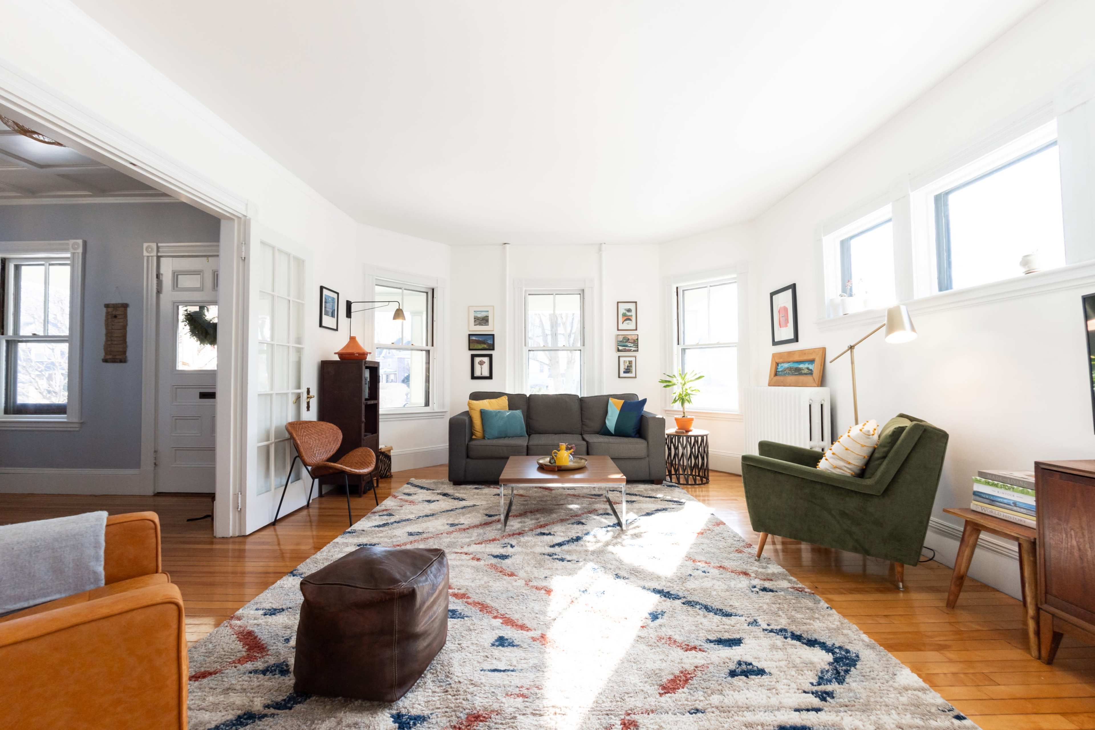 A bright living room features a gray sofa, green armchair, and a coffee table on a patterned rug, illuminated by natural light from multiple windows.