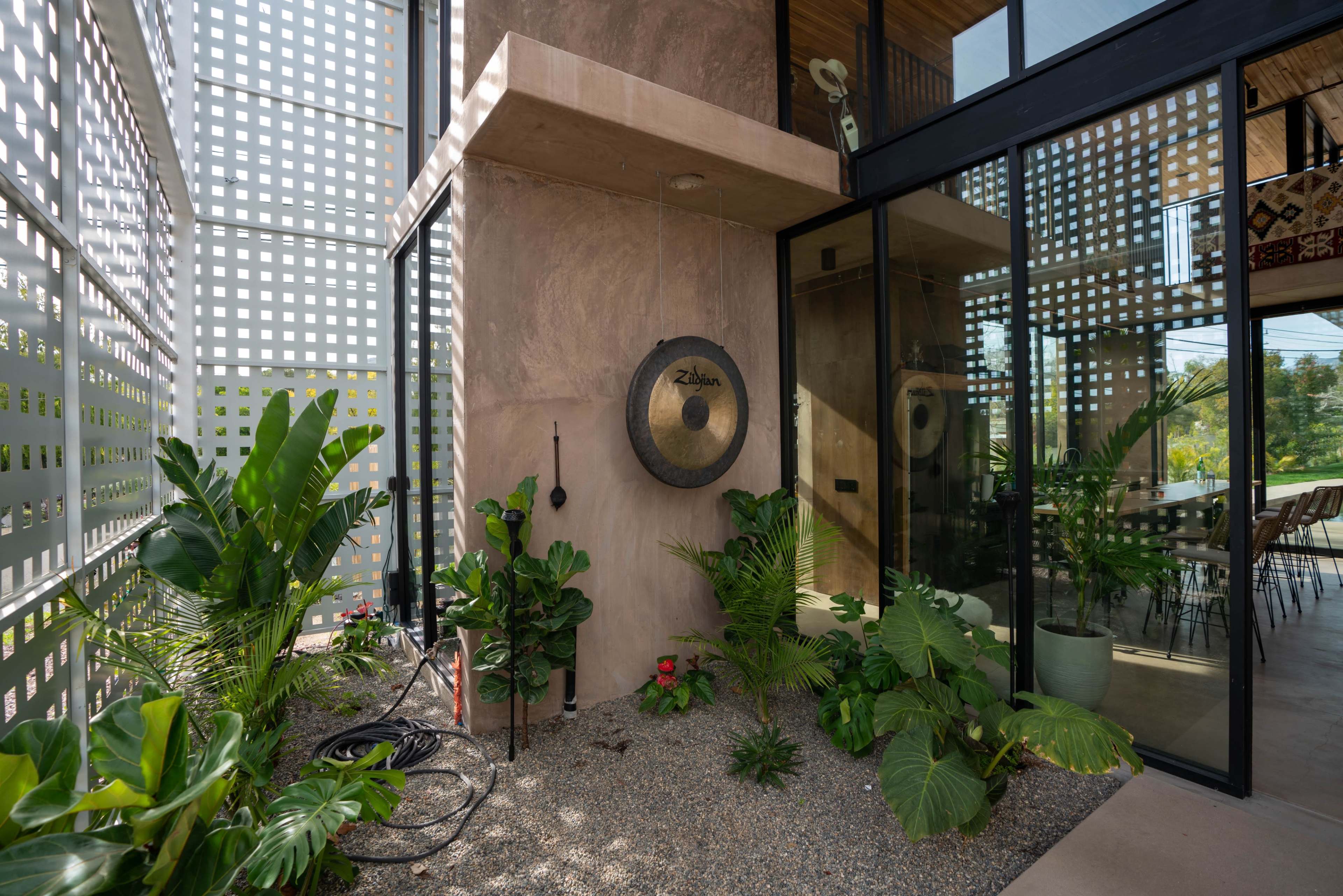 A modern entryway featuring a large gong mounted on a textured wall, surrounded by lush green plants and a gravel area.