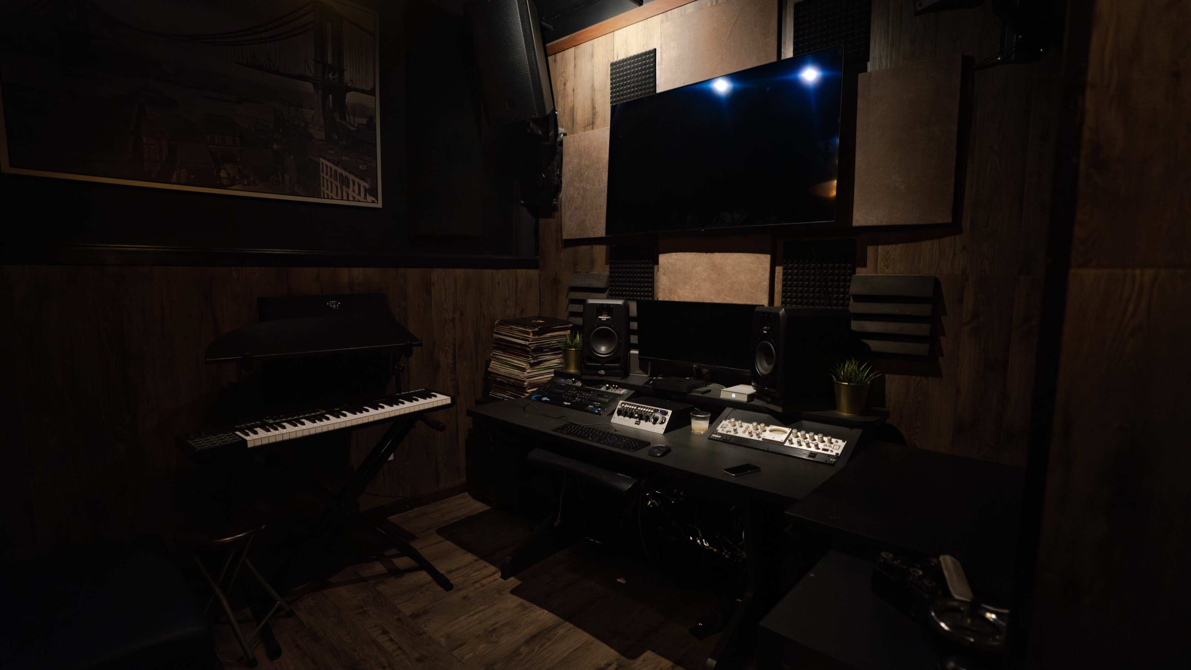 A music production studio features a keyboard, sound equipment, and a large screen mounted on the wall among wooden panels.