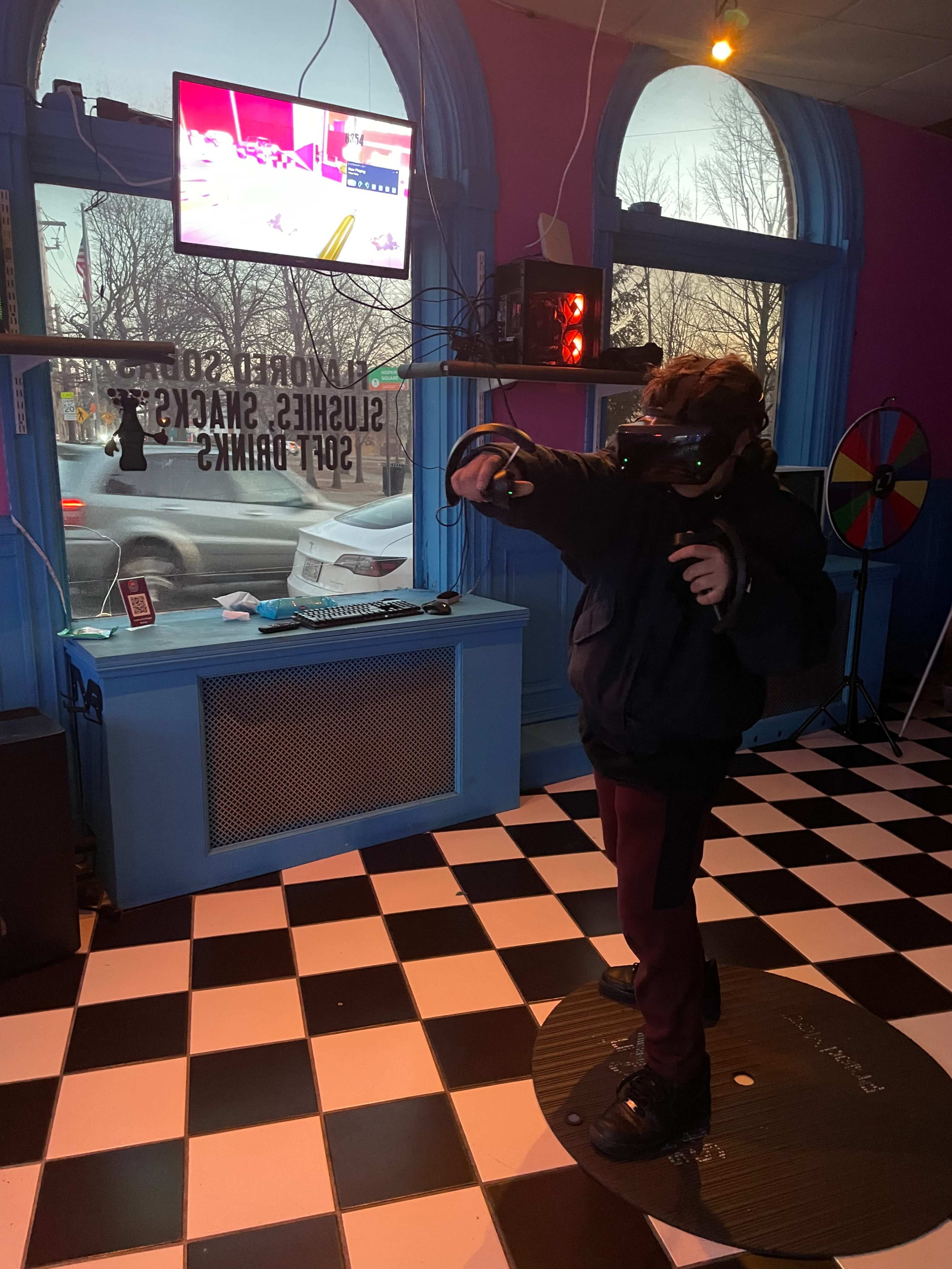 A child wearing a virtual reality headset stands on a circular platform, pointing while interacting with a display screen in a colorful room with checkered flooring.