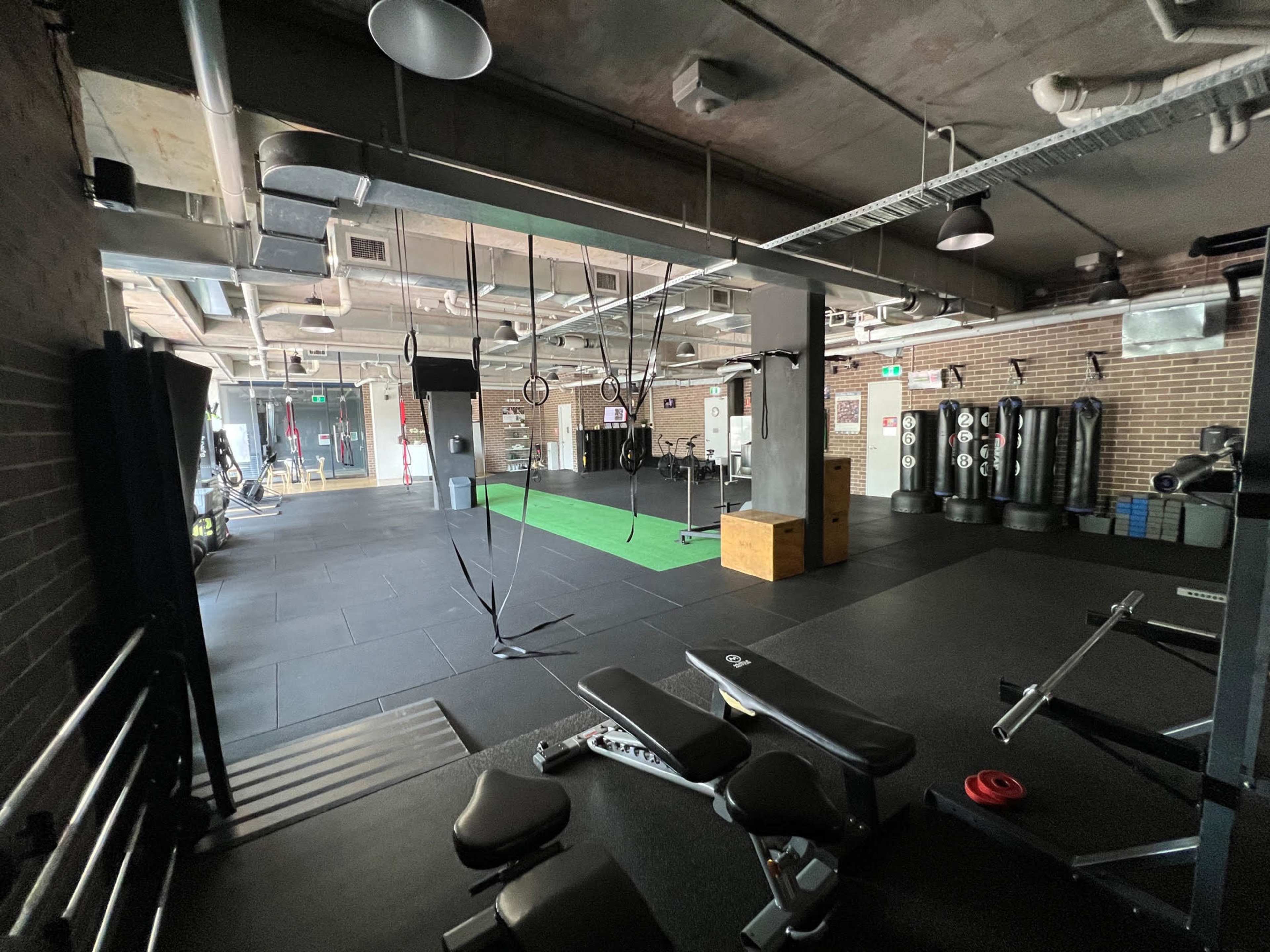 A spacious gym interior with various exercise equipment, including punching bags, a weight bench, and suspended training ropes, all set on black rubber flooring.