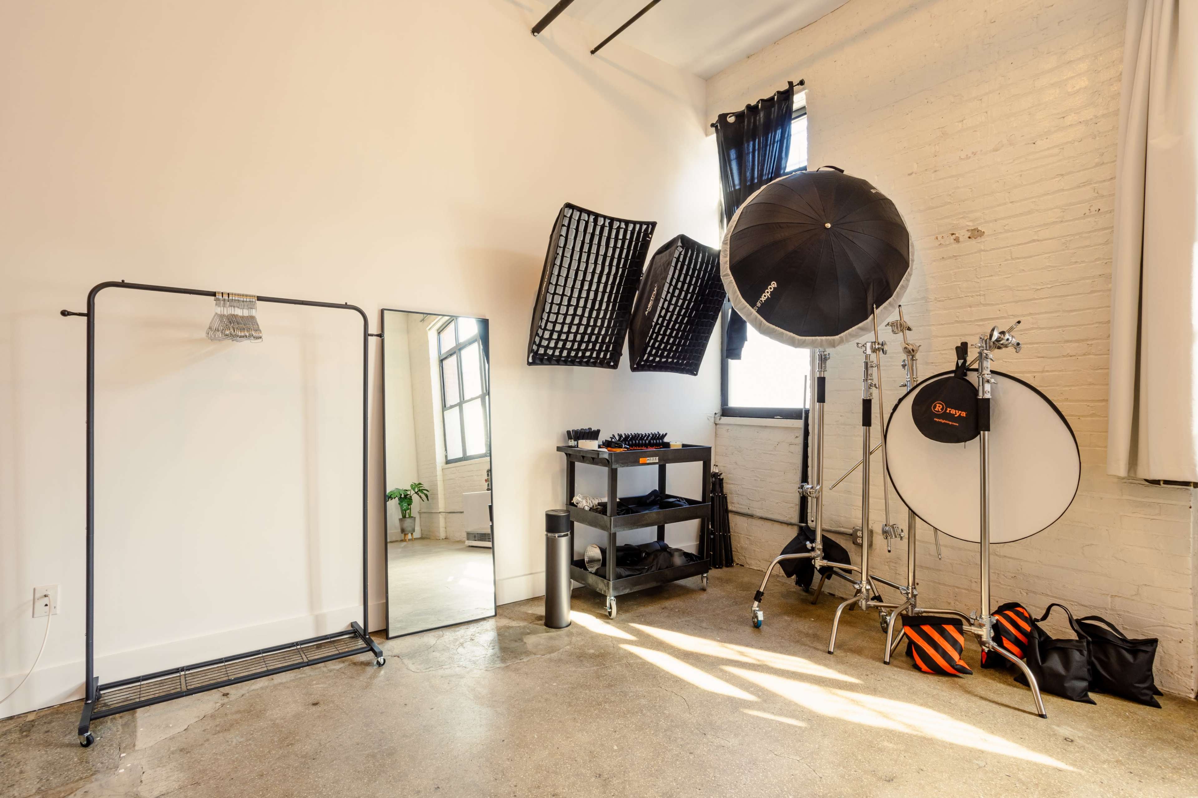 A photography studio features a large mirror, a clothing rack, various lighting equipment, and large windows letting in natural light.