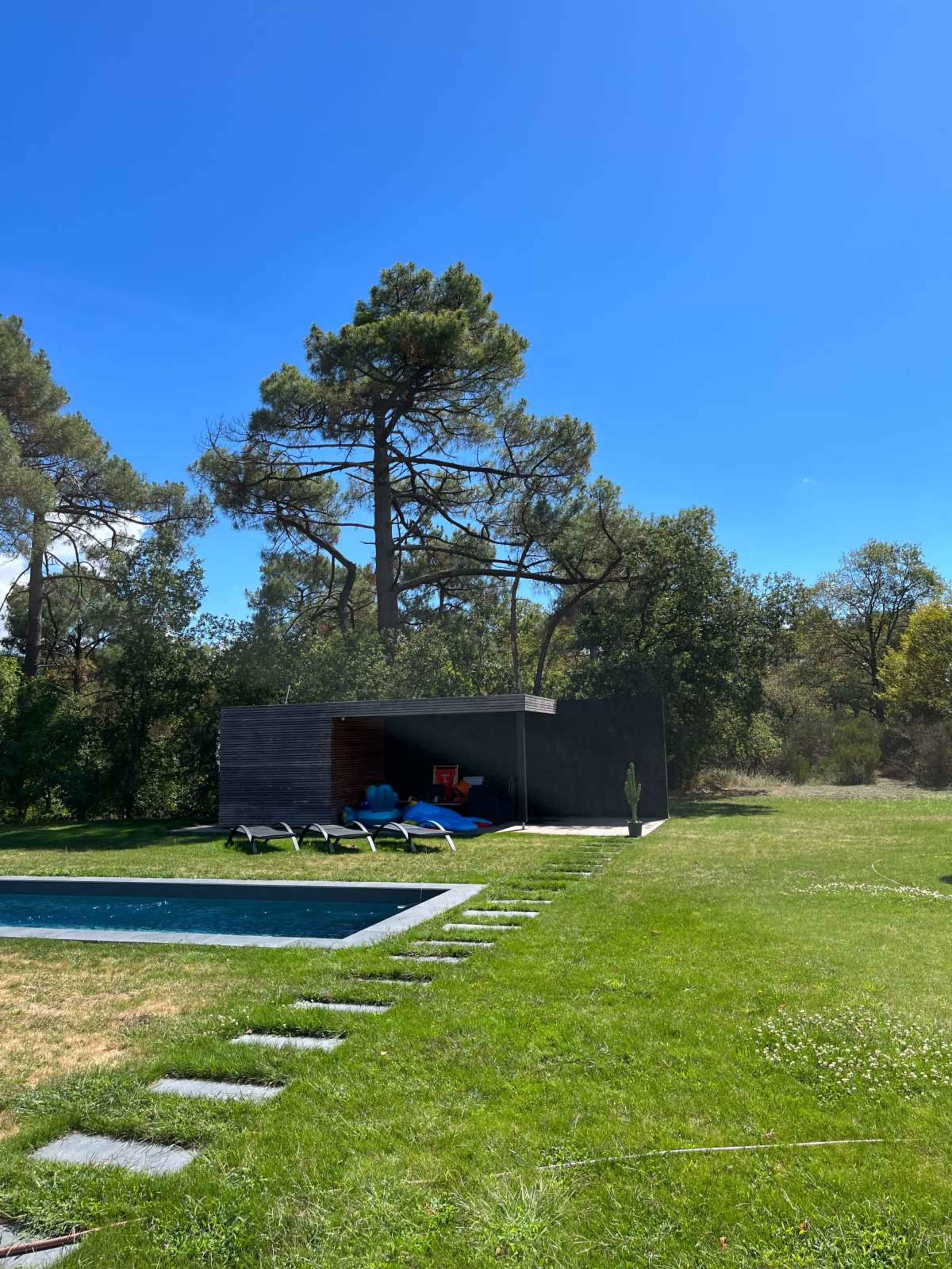 The image shows a modern poolside area with a rectangular swimming pool, lounge chairs, and a small structure set against a backdrop of trees and a clear blue sky.