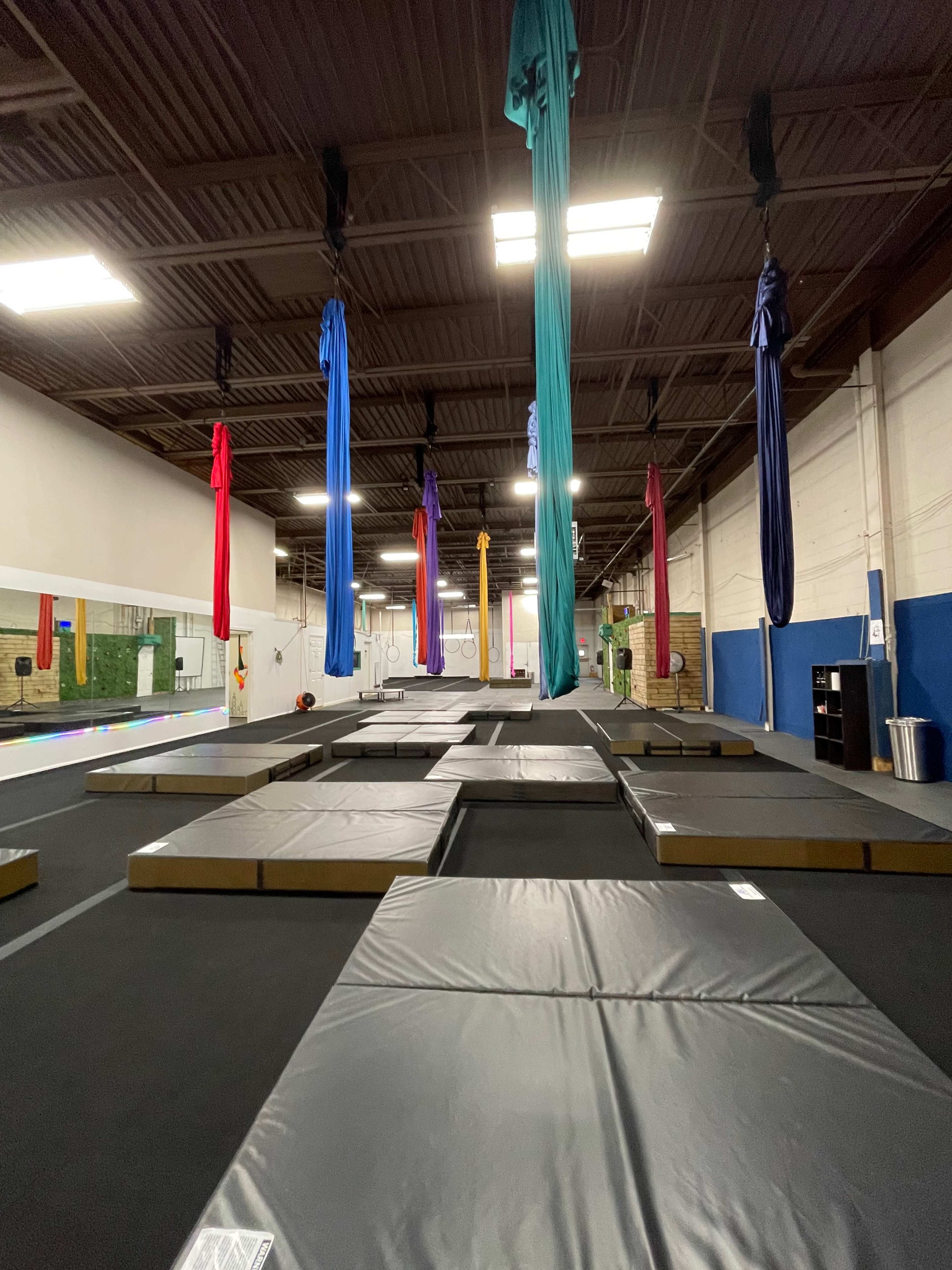 An indoor gym space with several large, padded mats and colorful aerial silks hanging from the ceiling.