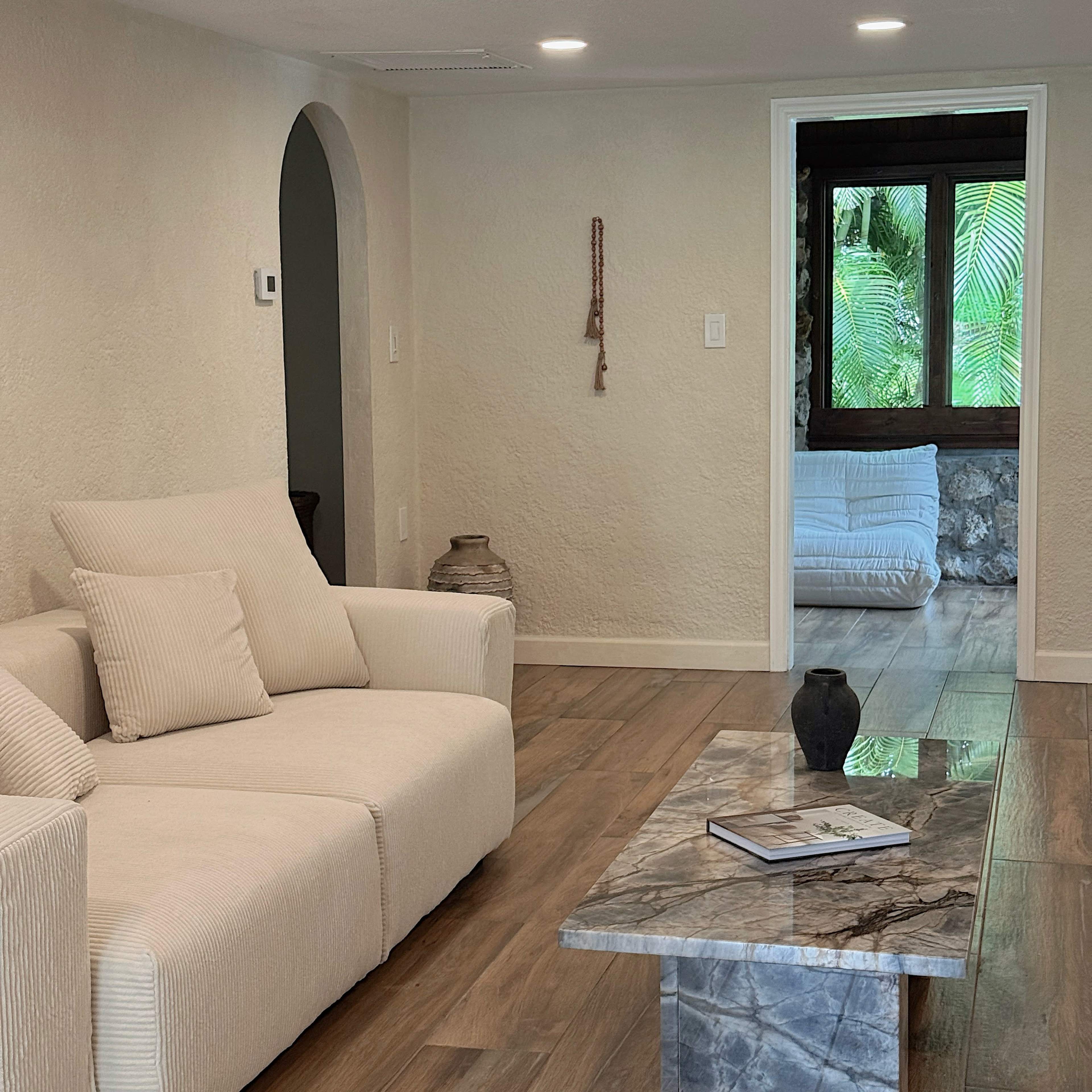 The image shows a modern, minimalist living room featuring a light-colored sofa, a marble coffee table, and a doorway leading to another room with visible greenery.