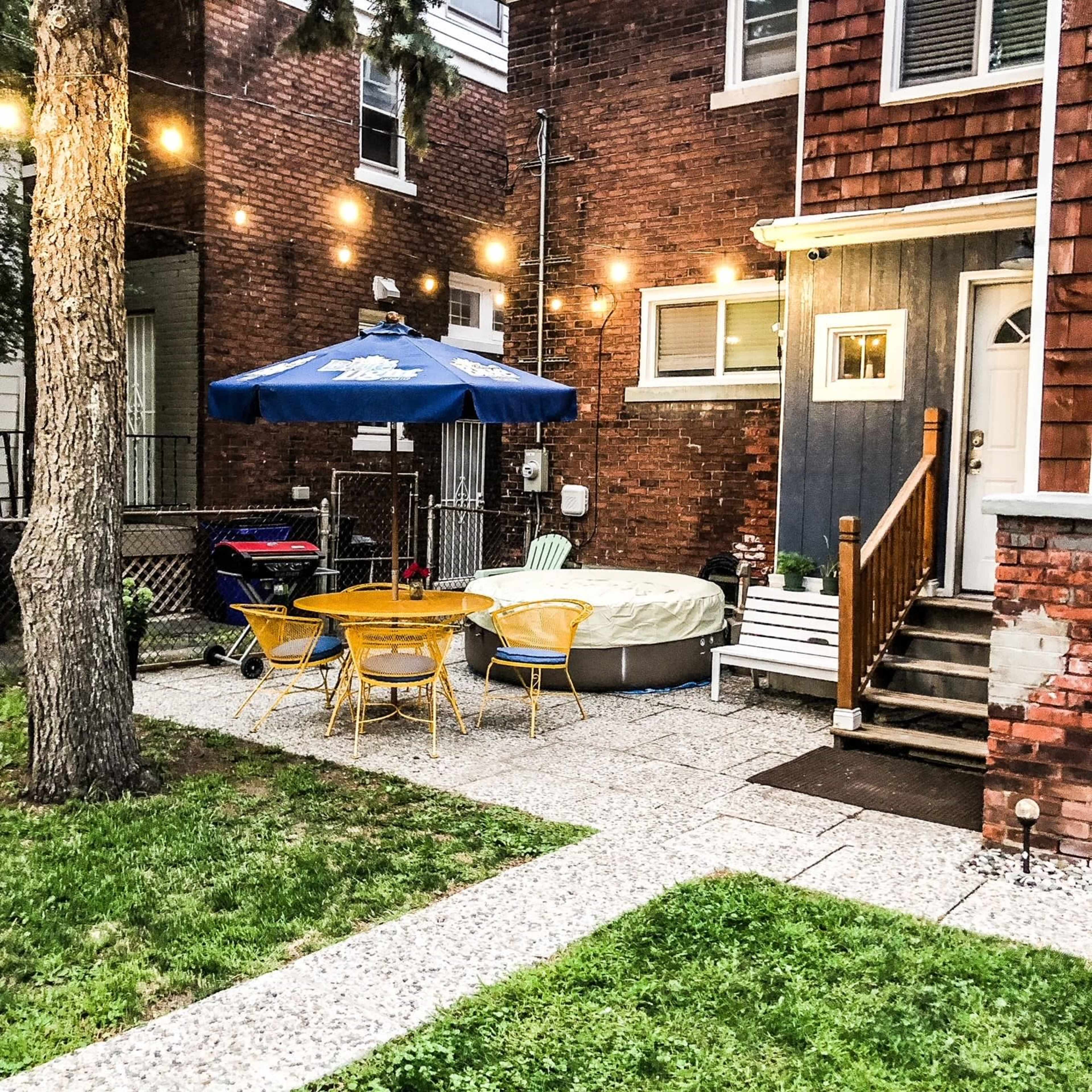 A backyard with a circular hot tub, a table with yellow chairs, a blue umbrella, and string lights hanging above.