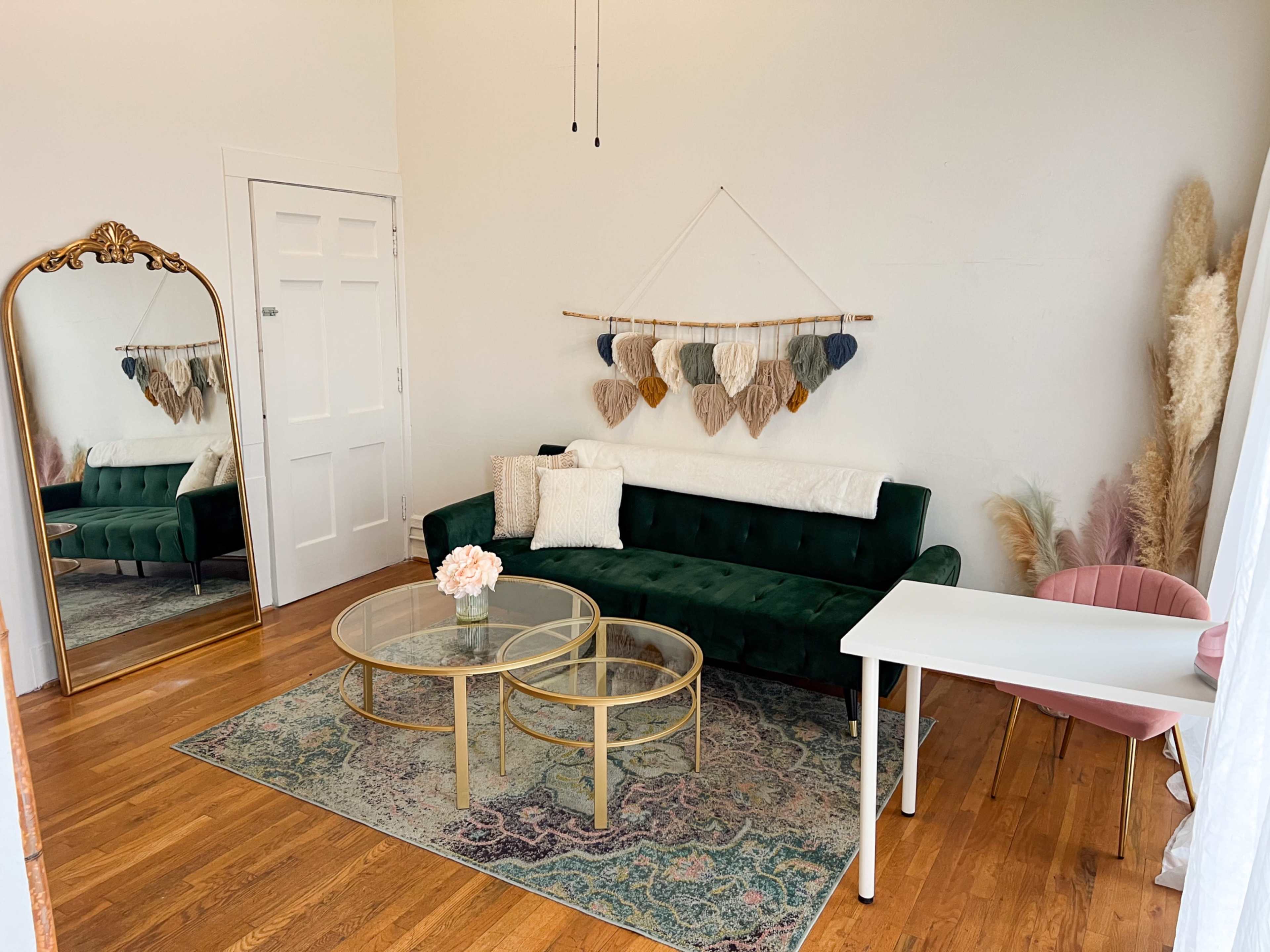 The image depicts a stylish living room with a green velvet sofa, a round coffee table, a pink chair, and a decorative mirror, alongside a wall adorned with hanging fabric hearts.