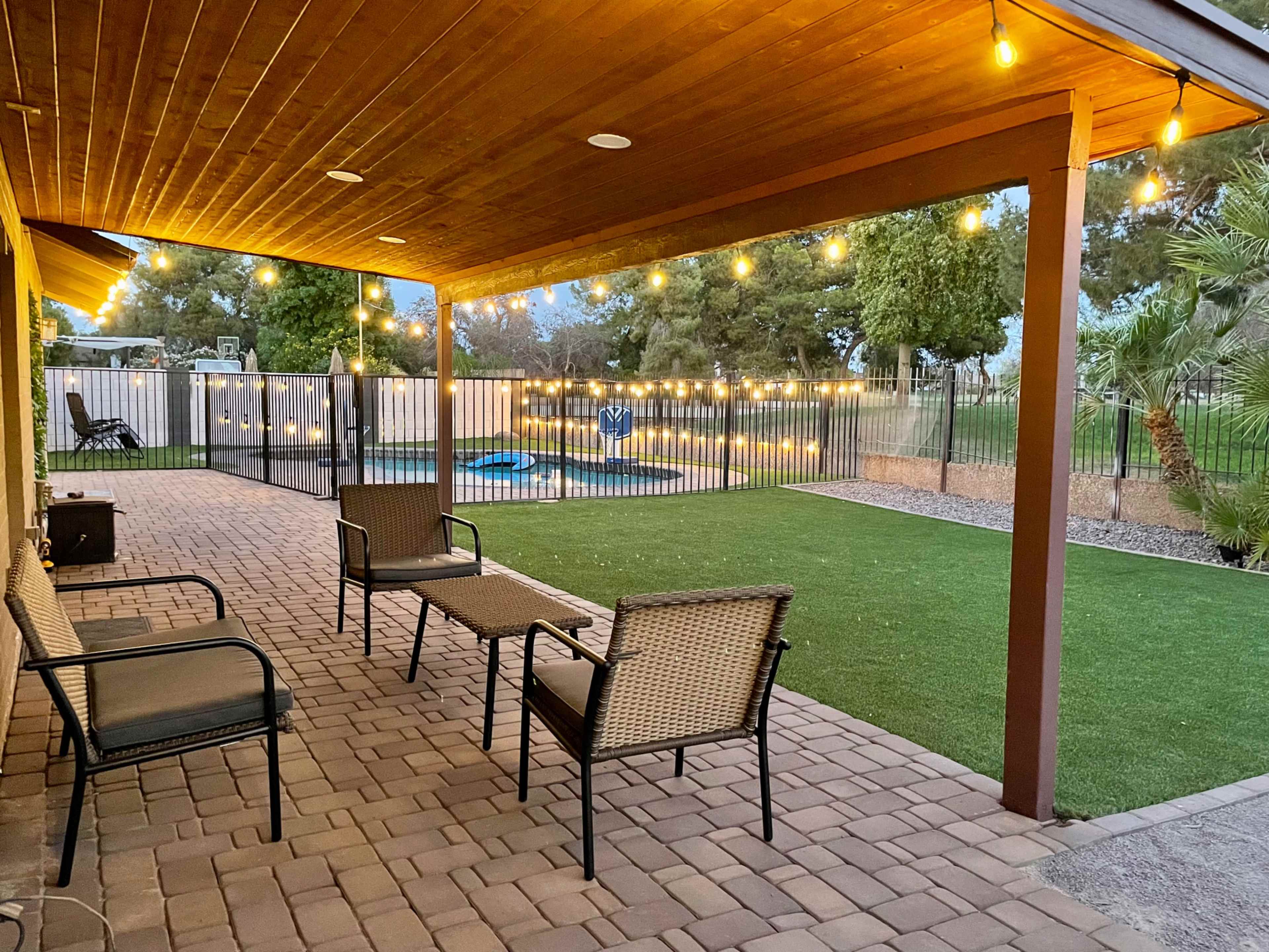 A covered patio features several chairs and a small table, illuminated by string lights, overlooking a fenced yard with a pool and greenery.
