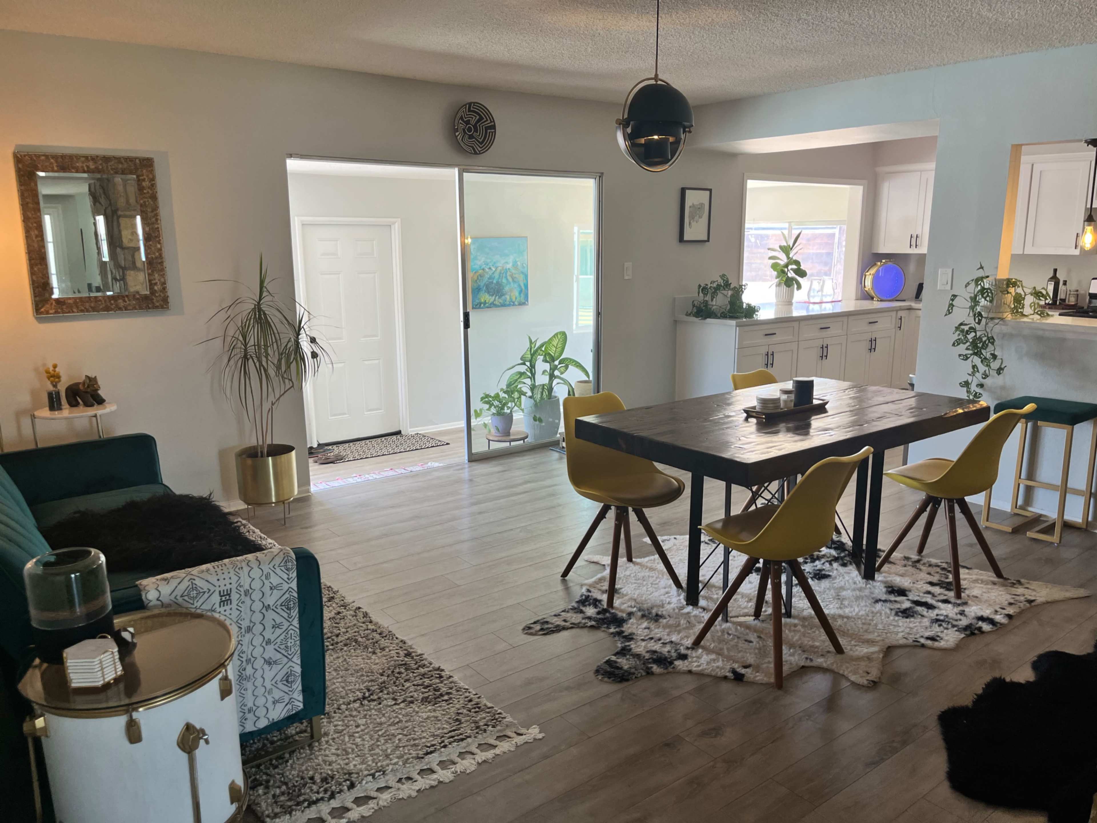 A modern living and dining area featuring a green sofa, a wooden dining table surrounded by yellow chairs, and plants throughout the space.