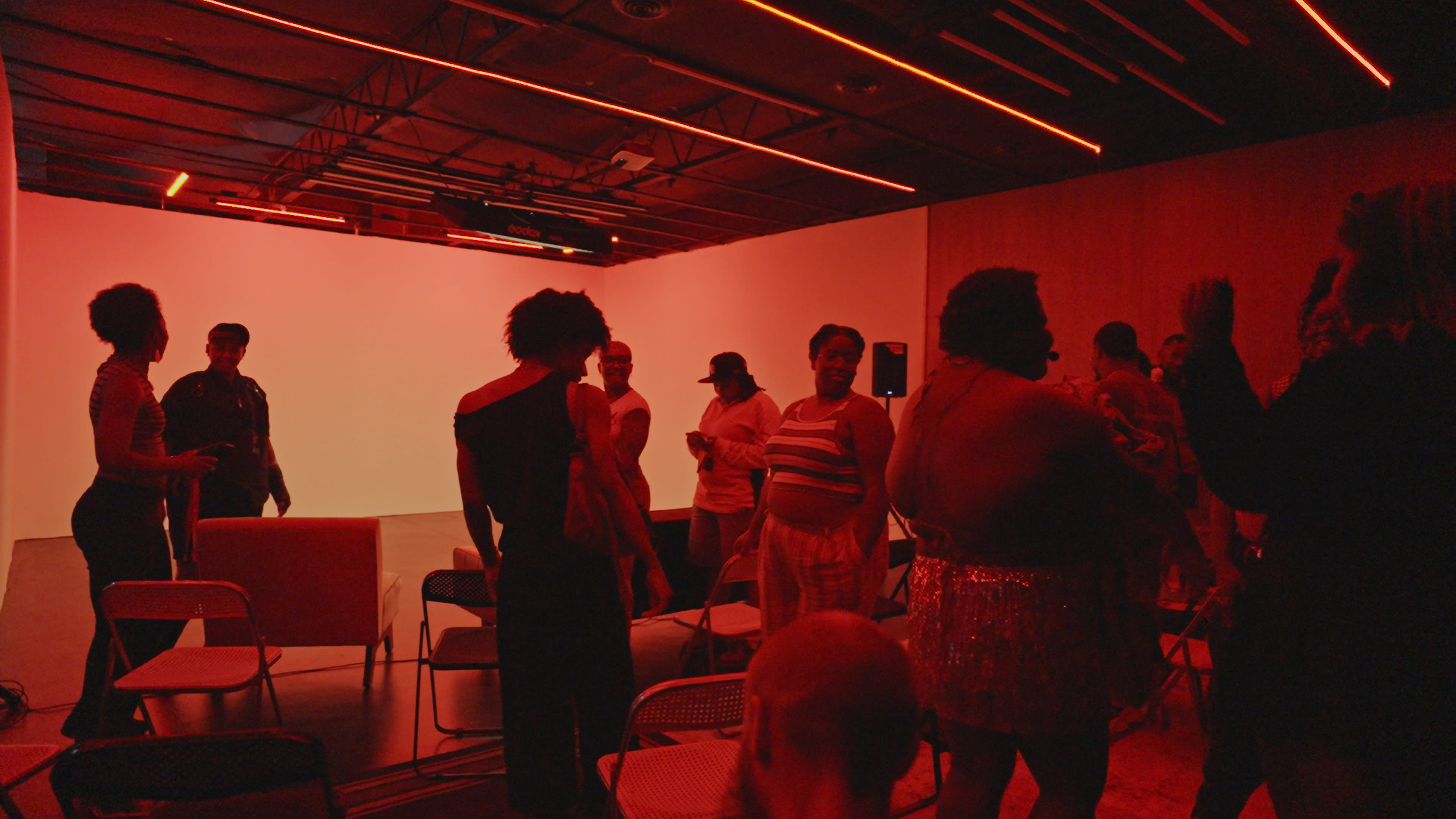 A group of people stands and mingles in a dimly lit room with red lighting and an empty white backdrop.