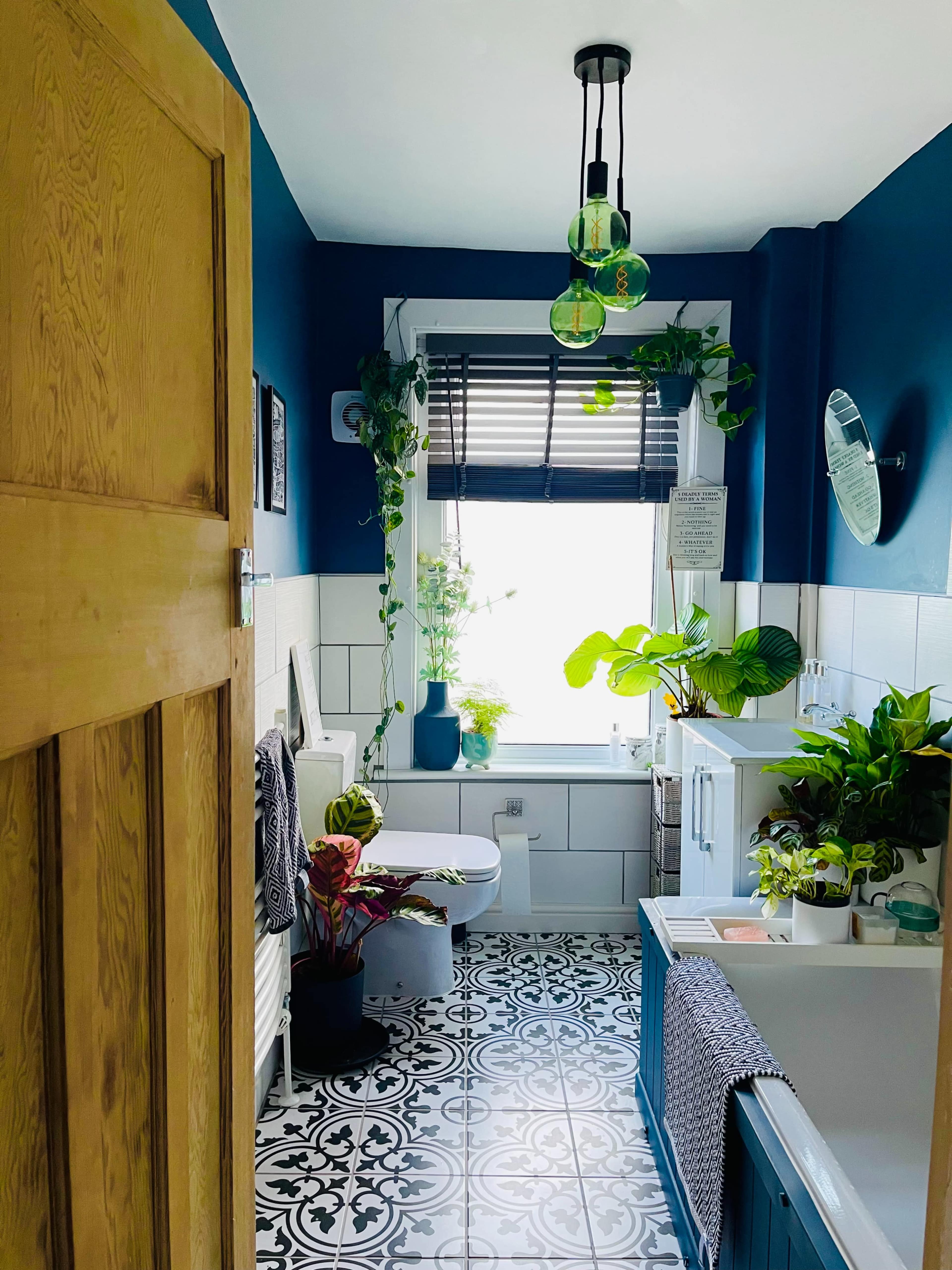 A modern bathroom features a bathtub, toilet, and sink, surrounded by plants and decorated with blue walls and patterned tiles.