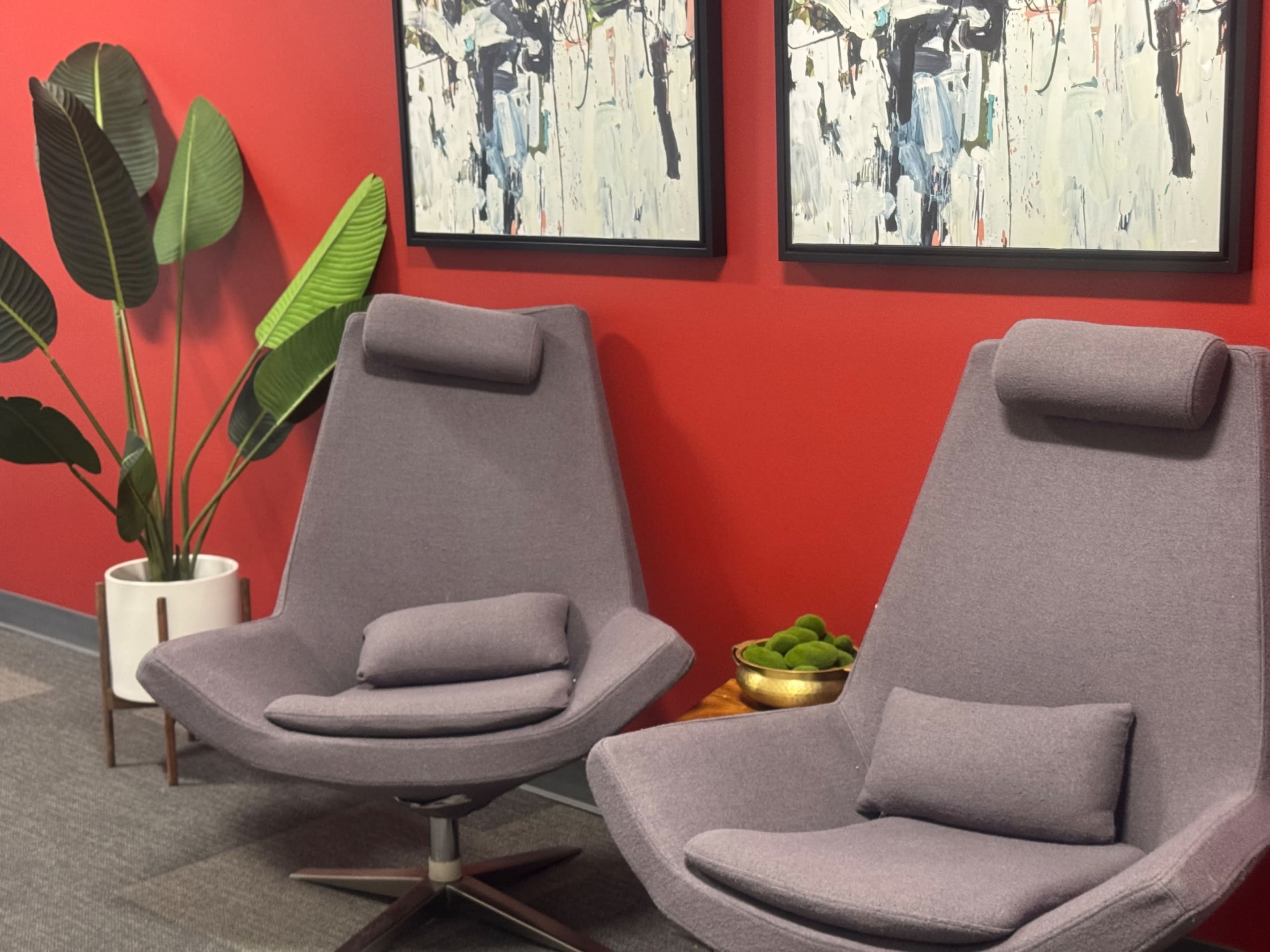 The image shows two gray chairs with headrests positioned in front of a red wall, with two abstract paintings above them and a potted plant nearby.