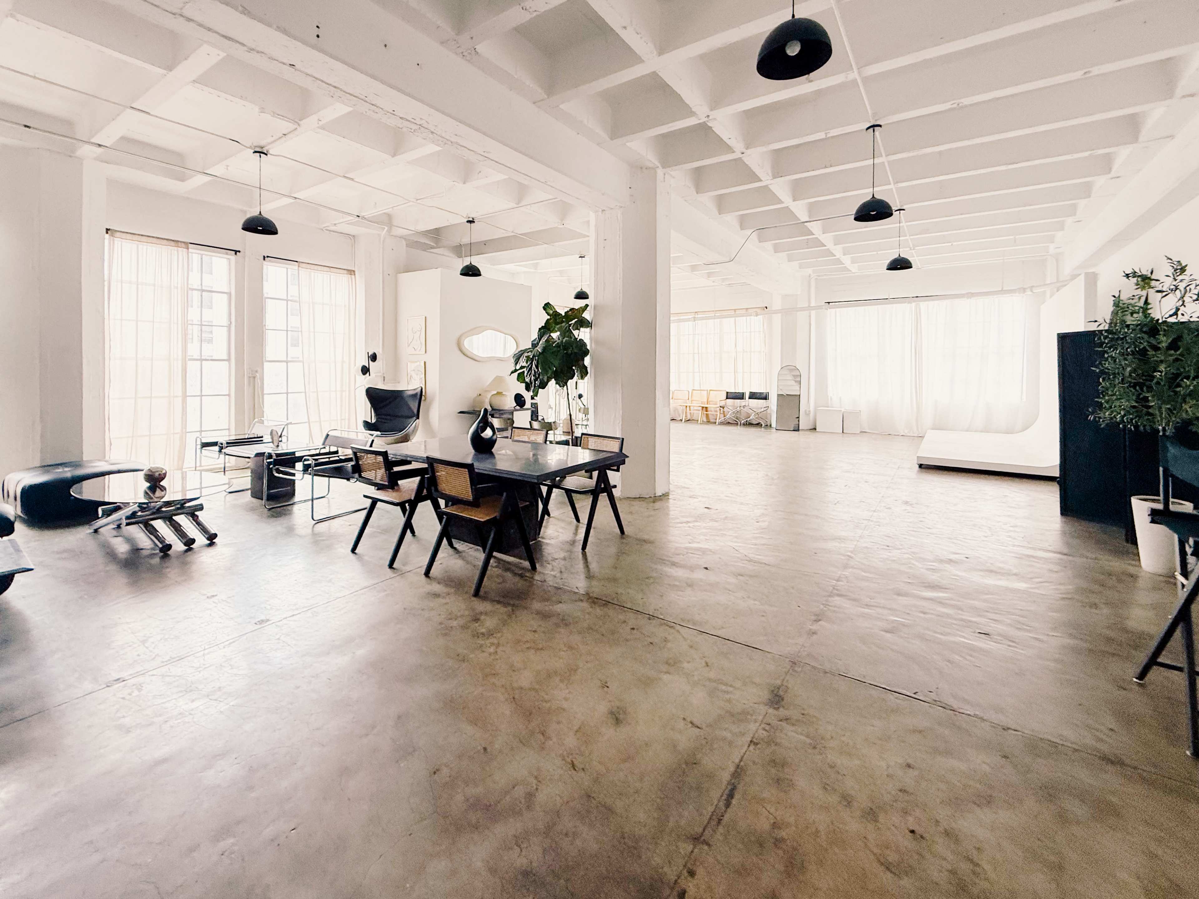 The image shows a spacious, minimalistic interior filled with natural light, featuring a dining table with chairs, a lounge area, and large windows.