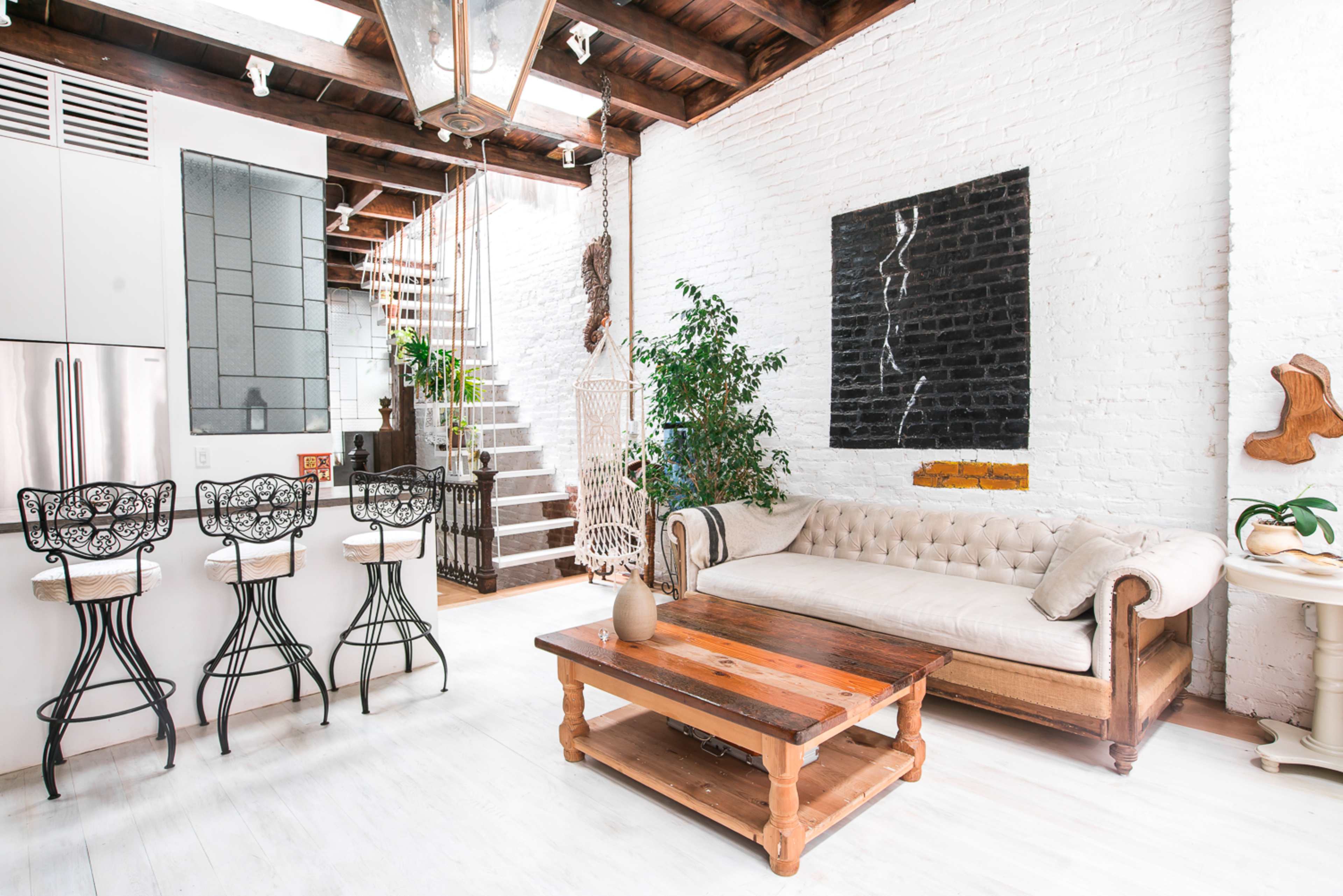 A modern living space features a white brick wall, a beige sofa, a wooden coffee table, and bar stools at a kitchen counter, with a staircase in the background leading to an upper level.