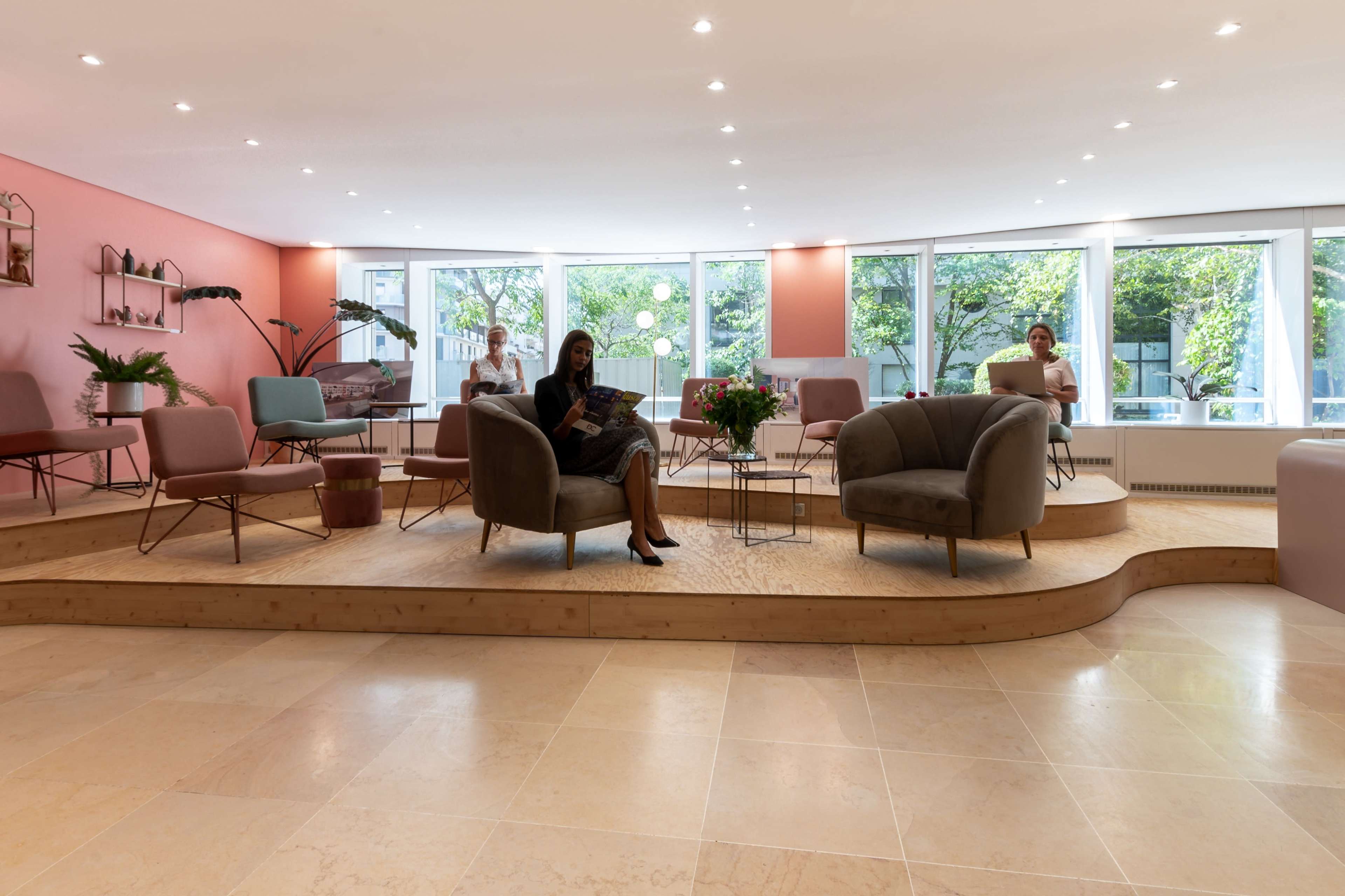 A modern indoor lounge area with a woman seated on a raised platform, surrounded by various seating arrangements and large windows displaying greenery outside.