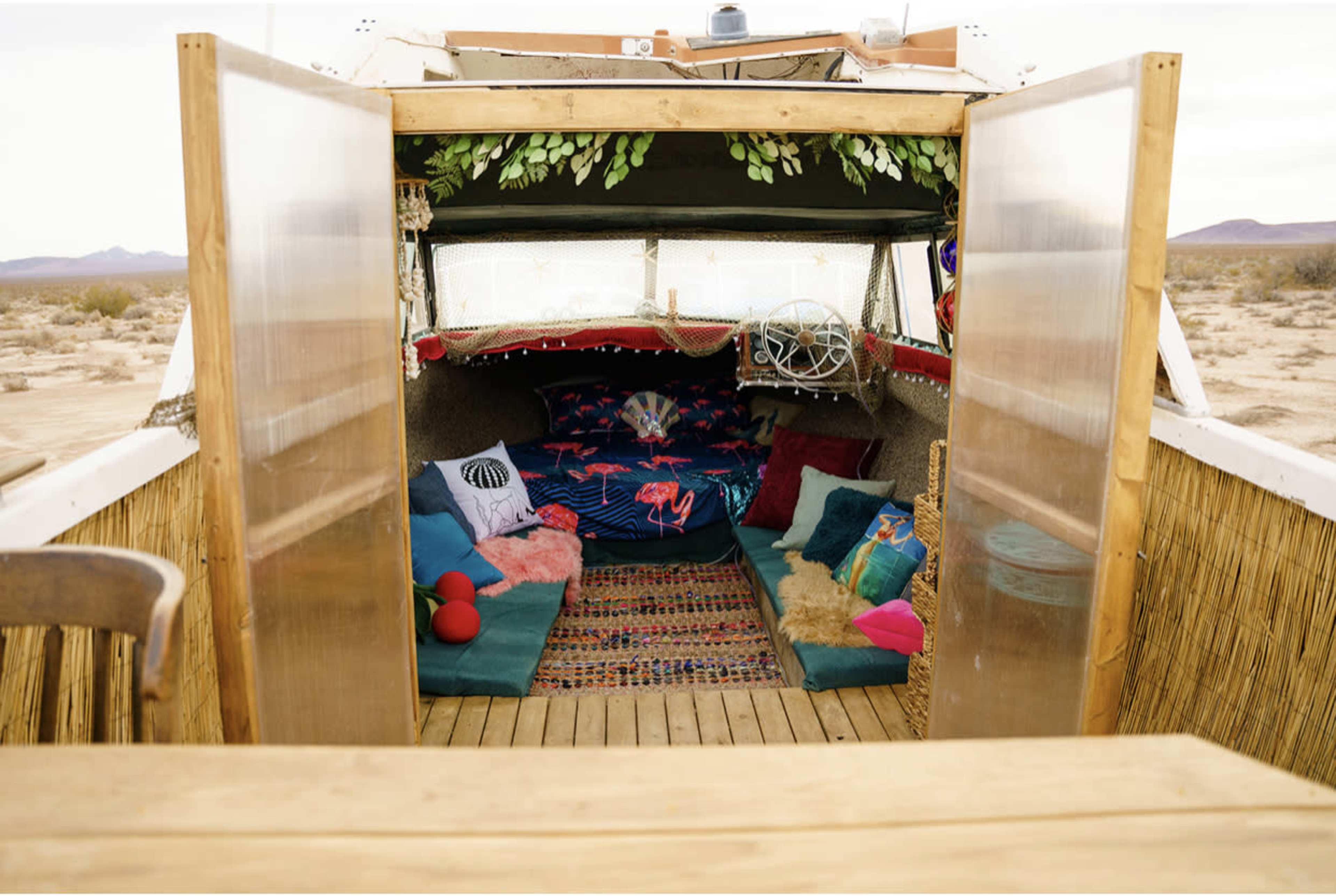 The image shows the interior of a rustic camper, featuring colorful cushions and a woven rug, set in a desert landscape.