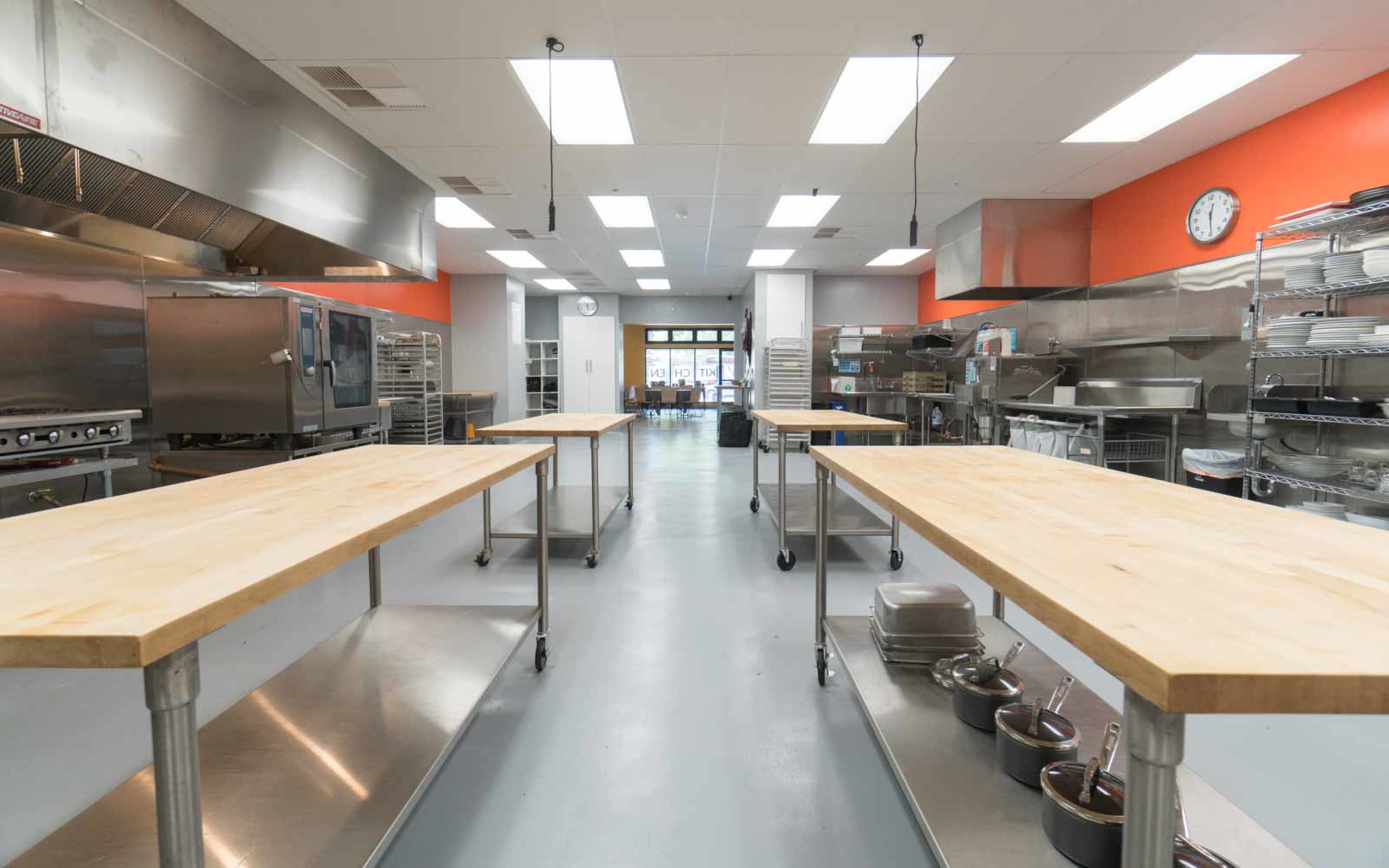 A bright commercial kitchen with stainless steel appliances and wooden worktables arranged in a spacious layout.