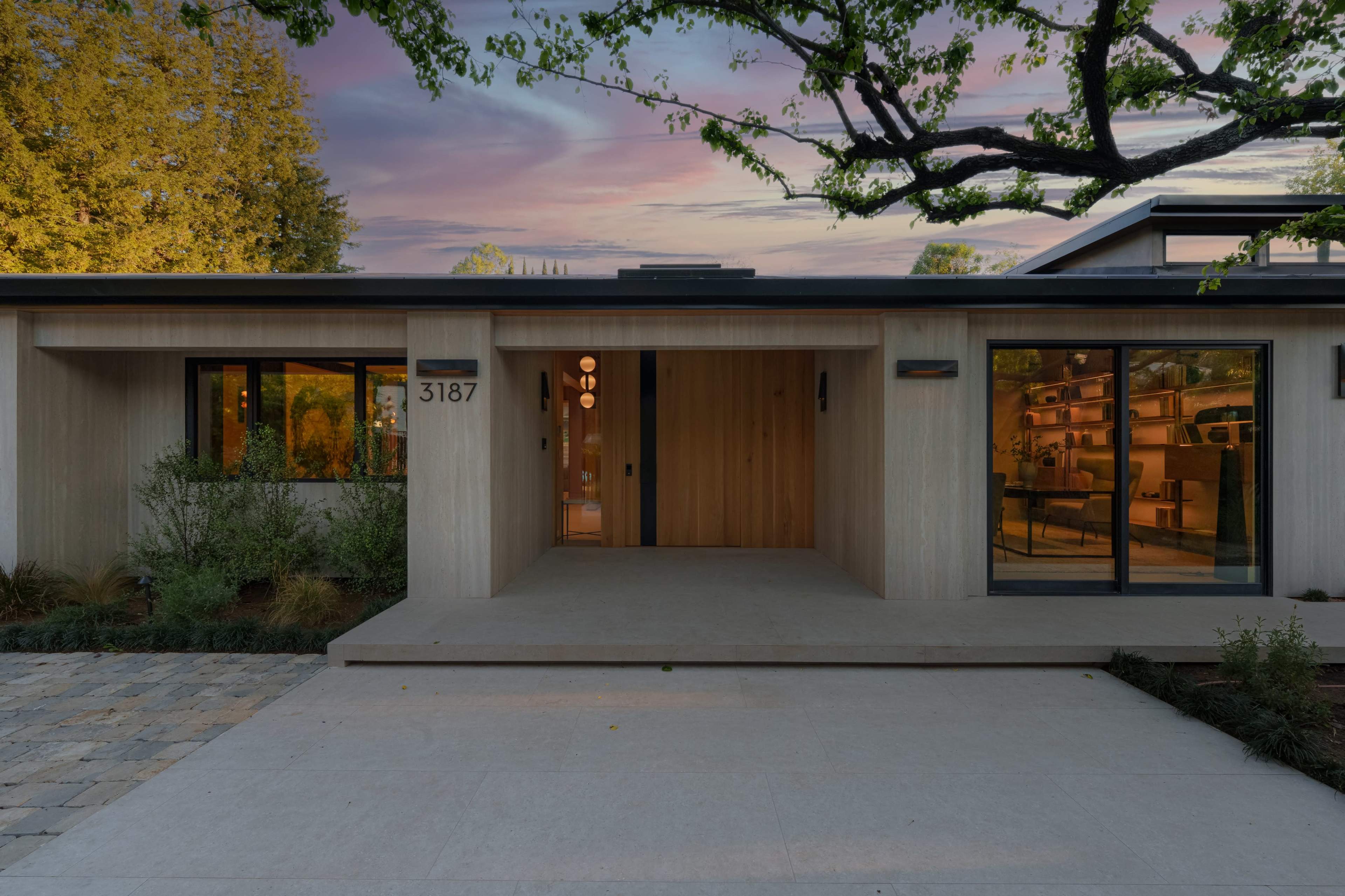 The image shows a modern house with wooden paneling, large windows, and a concrete pathway leading to its entrance.