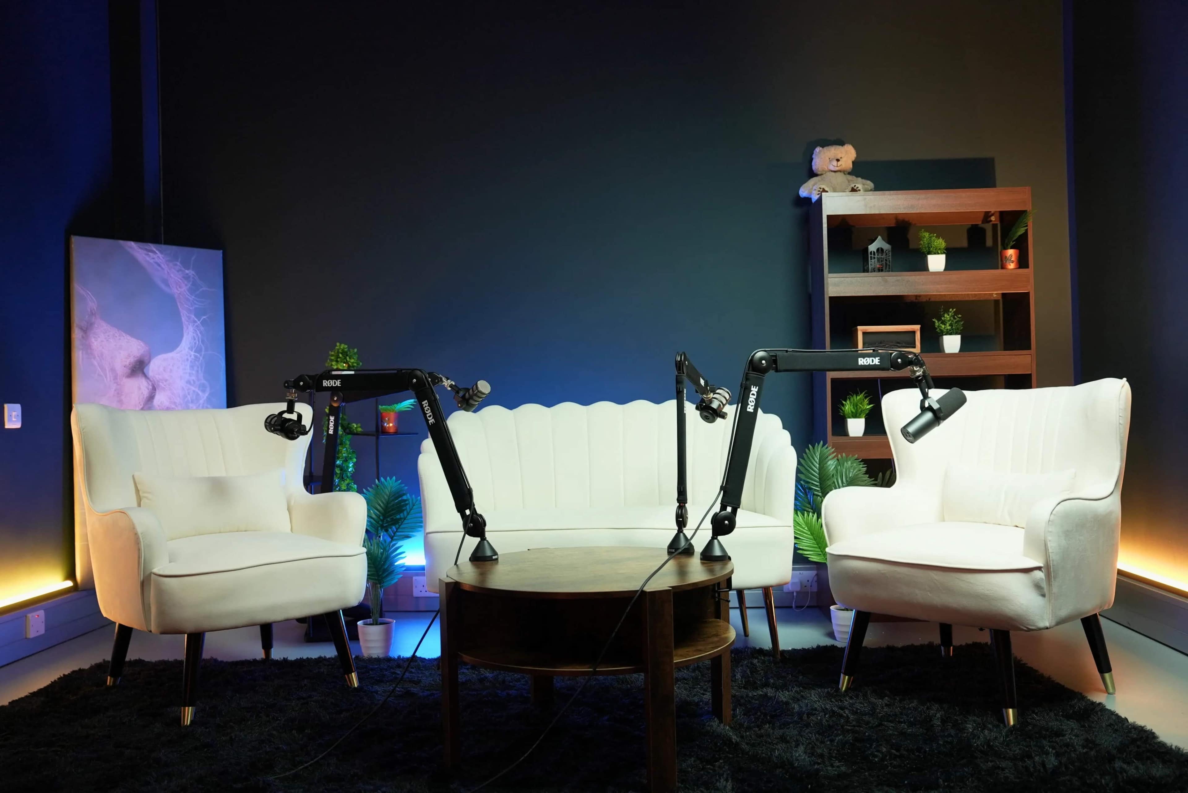 The image shows a modern podcast or interview set featuring two white armchairs, a round wooden table, and microphone stands, all arranged against a dark wall with decorative shelves and plants.
