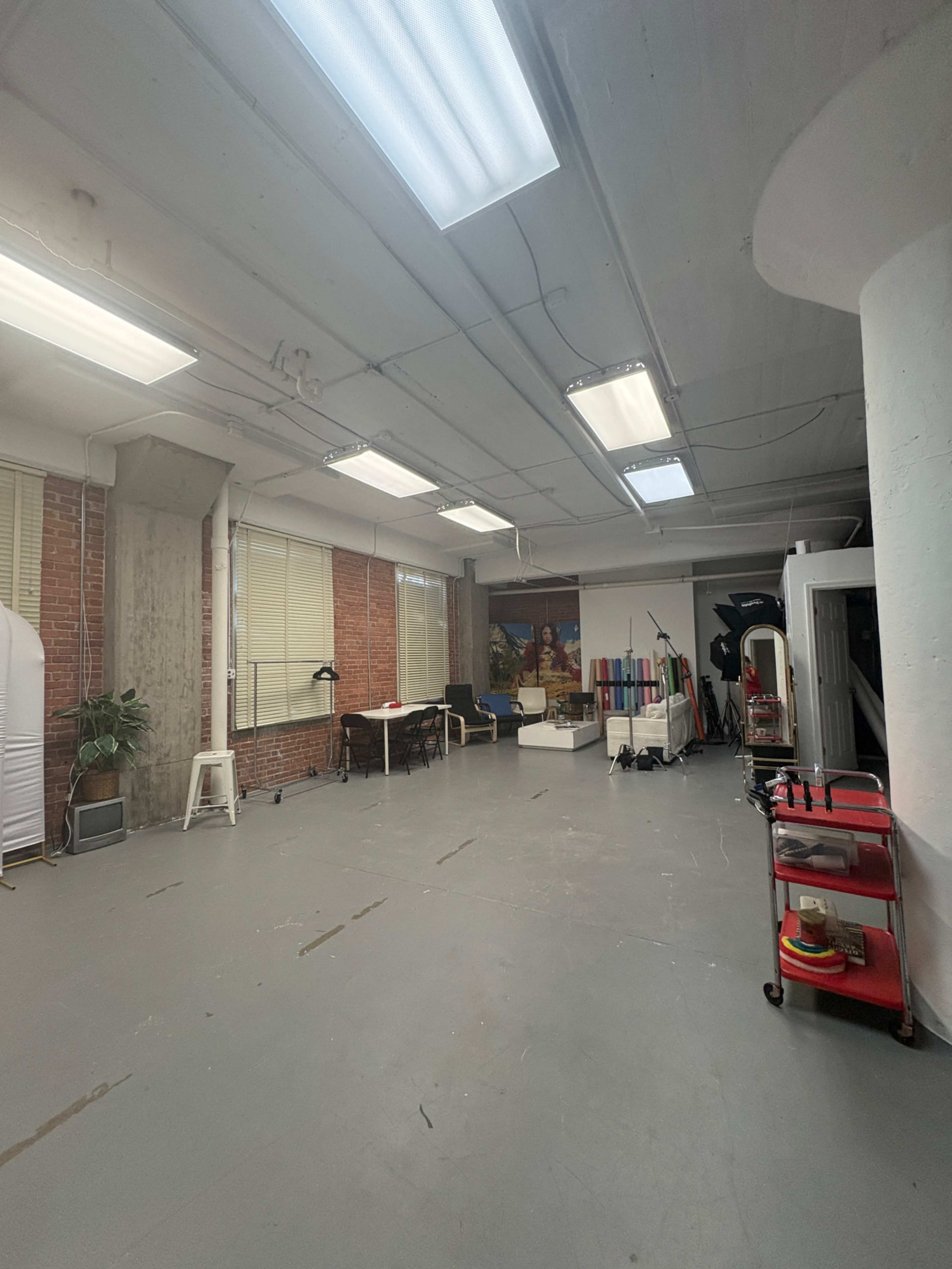 The image shows a spacious, industrial-style room with concrete walls, bright overhead lights, and various furniture and equipment along the sides.