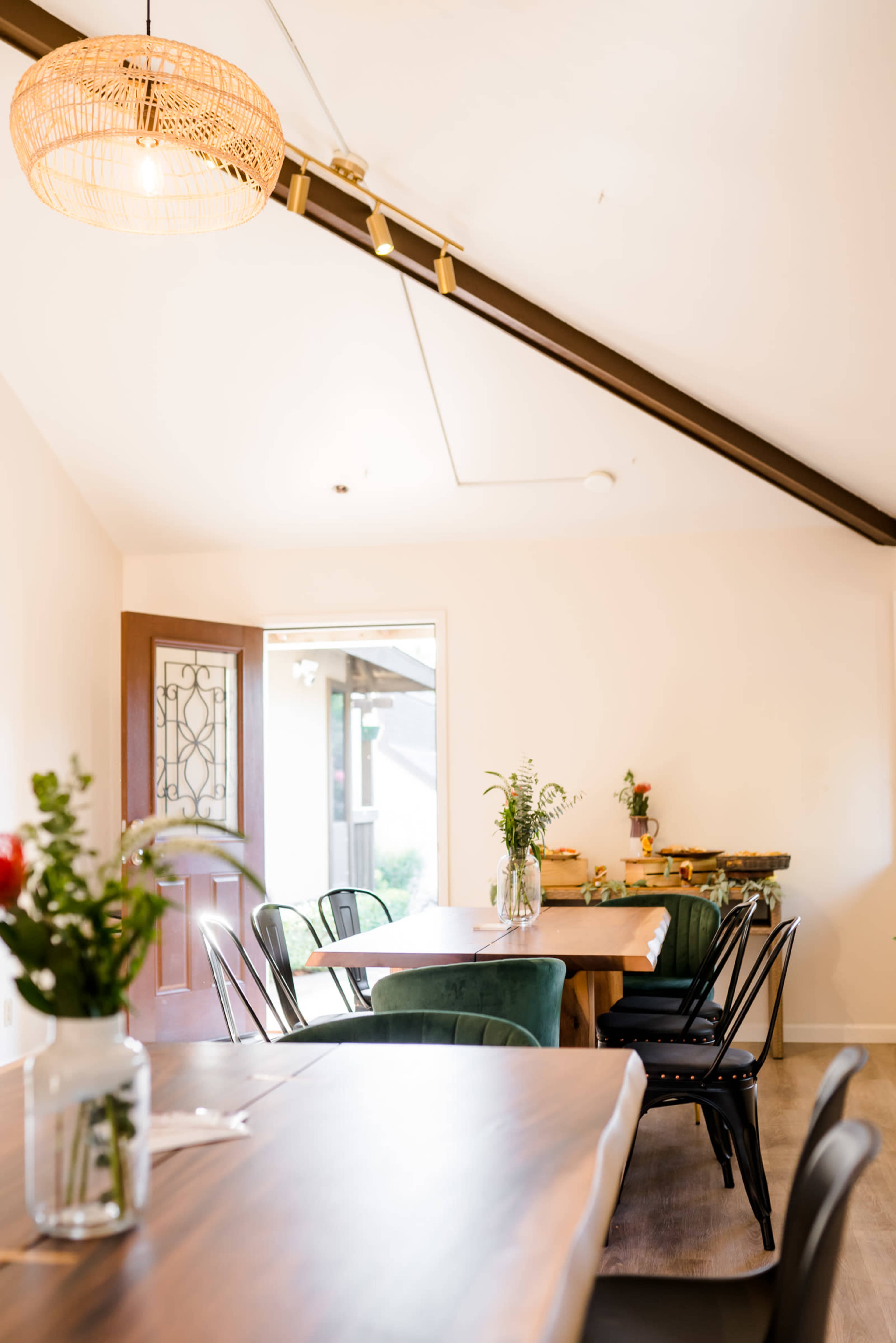 A well-lit dining area features a wooden table surrounded by chairs, with a side table displaying plants and flowers.
