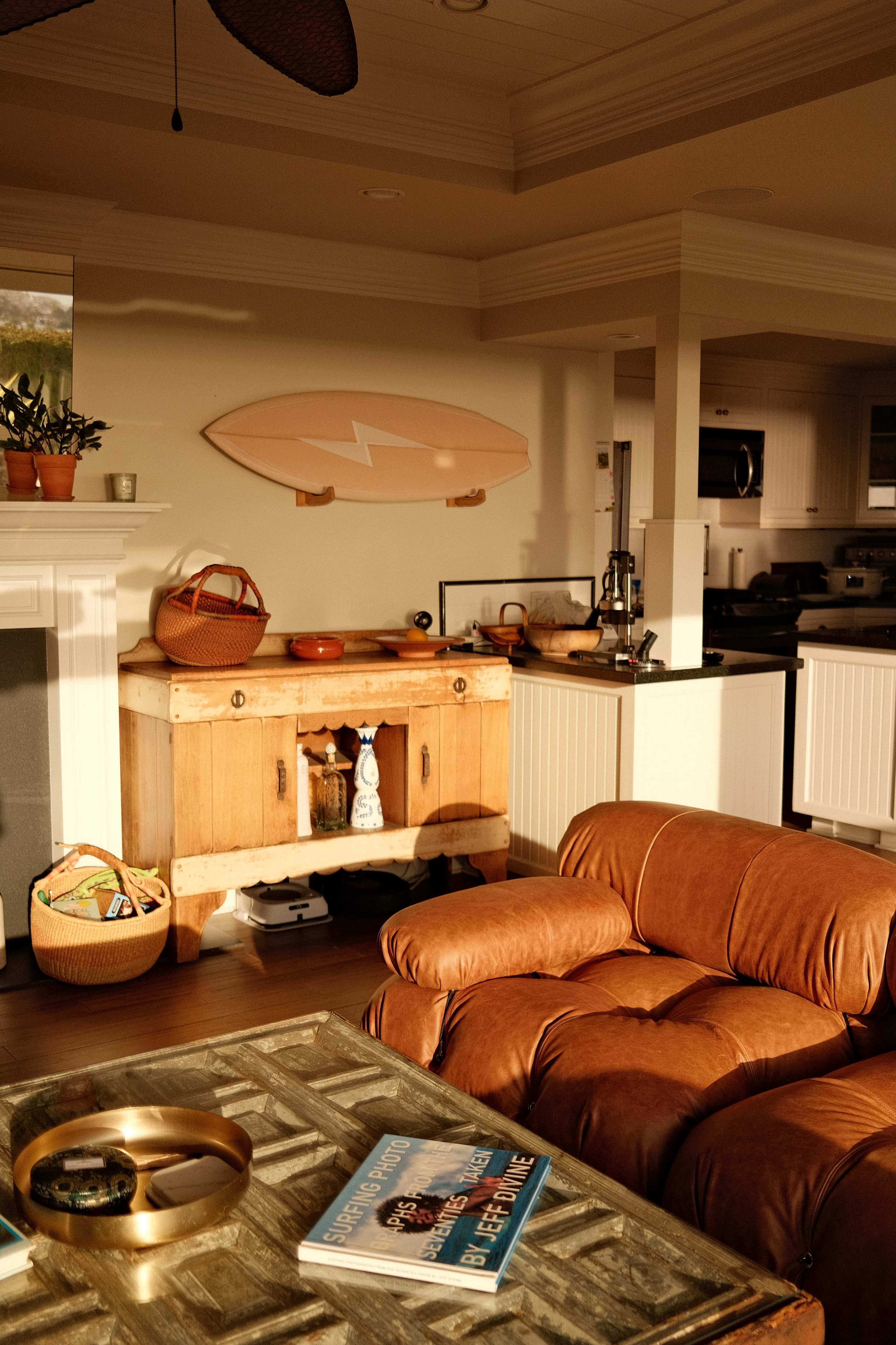 The image shows a cozy living space with a leather chair, a wooden cabinet, a surfboard on the wall, and various decorative items scattered around.