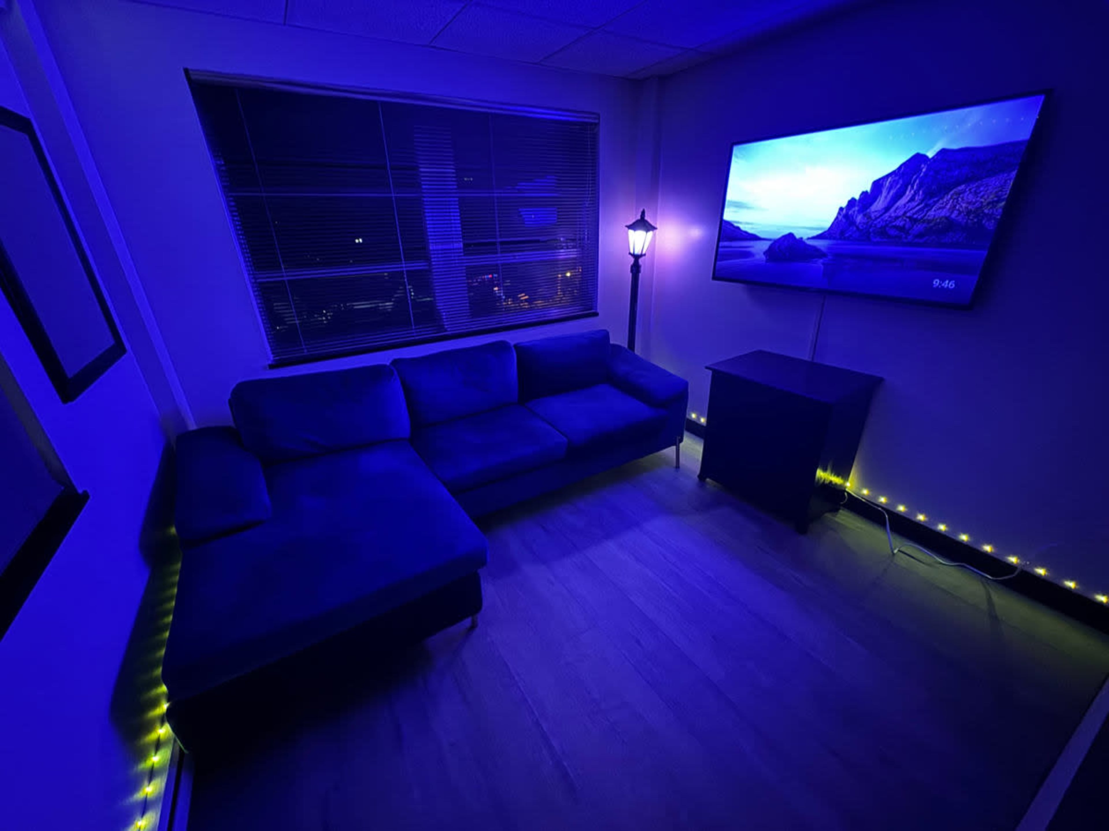 A cozy room features a blue sectional sofa, a television mounted on the wall, and soft LED lighting along the floor.