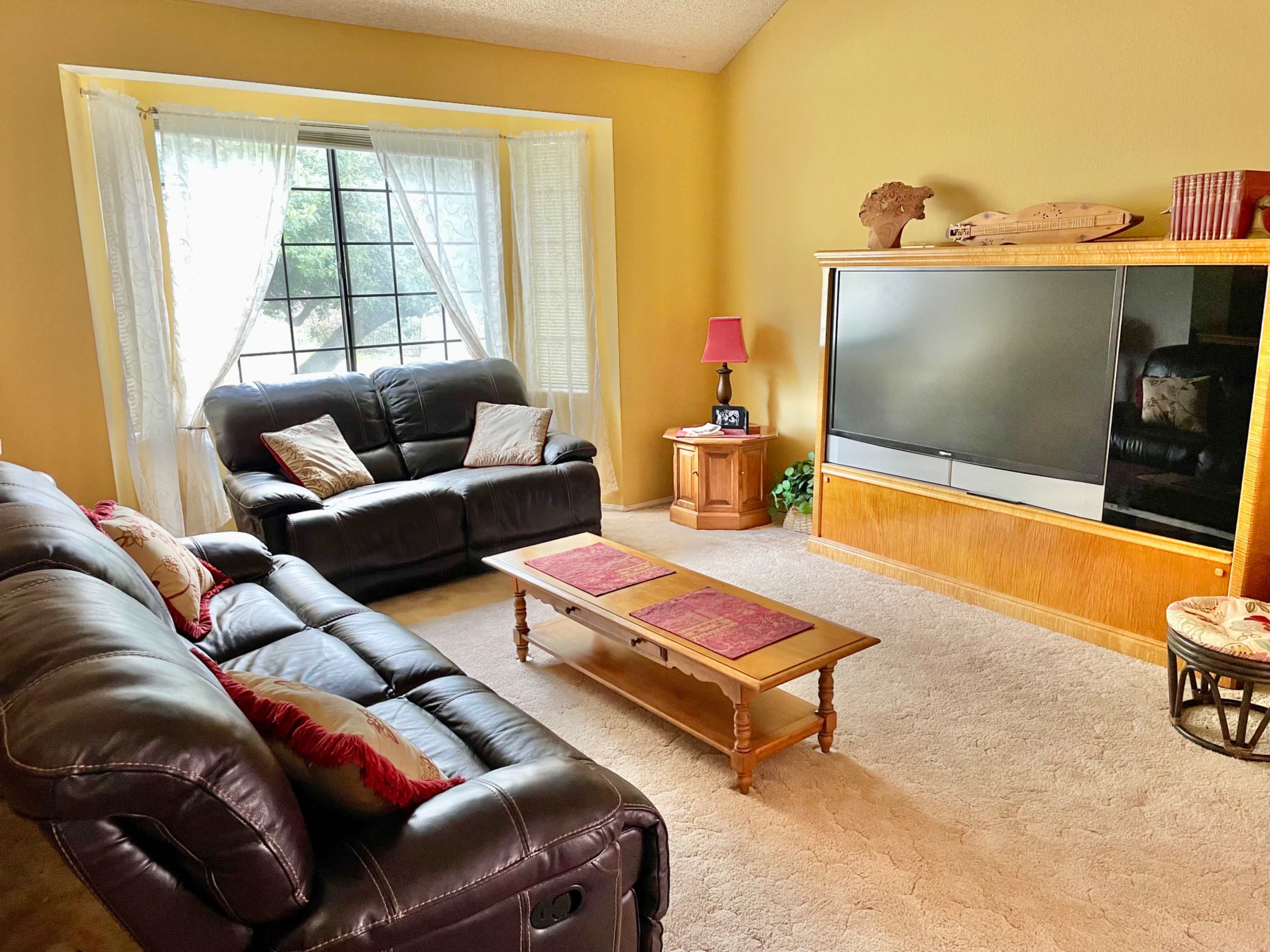 The image shows a living room with a large window, two black leather sofas, a wooden coffee table with decorative items, and a television mounted on an oak cabinet.