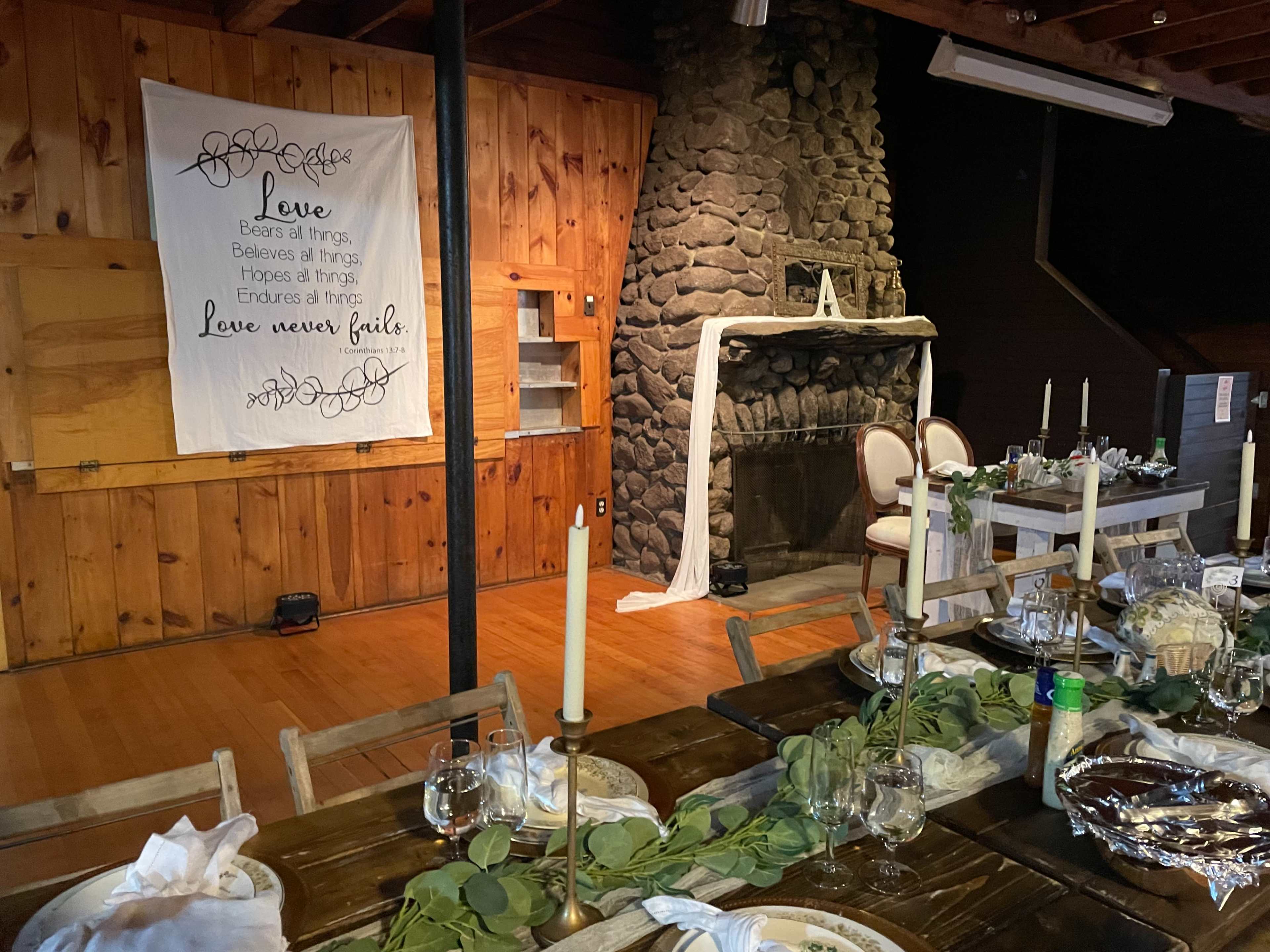 A wooden interior with a stone fireplace and a dining table set with candles and greenery, alongside a large banner that reads "Love bears all things."