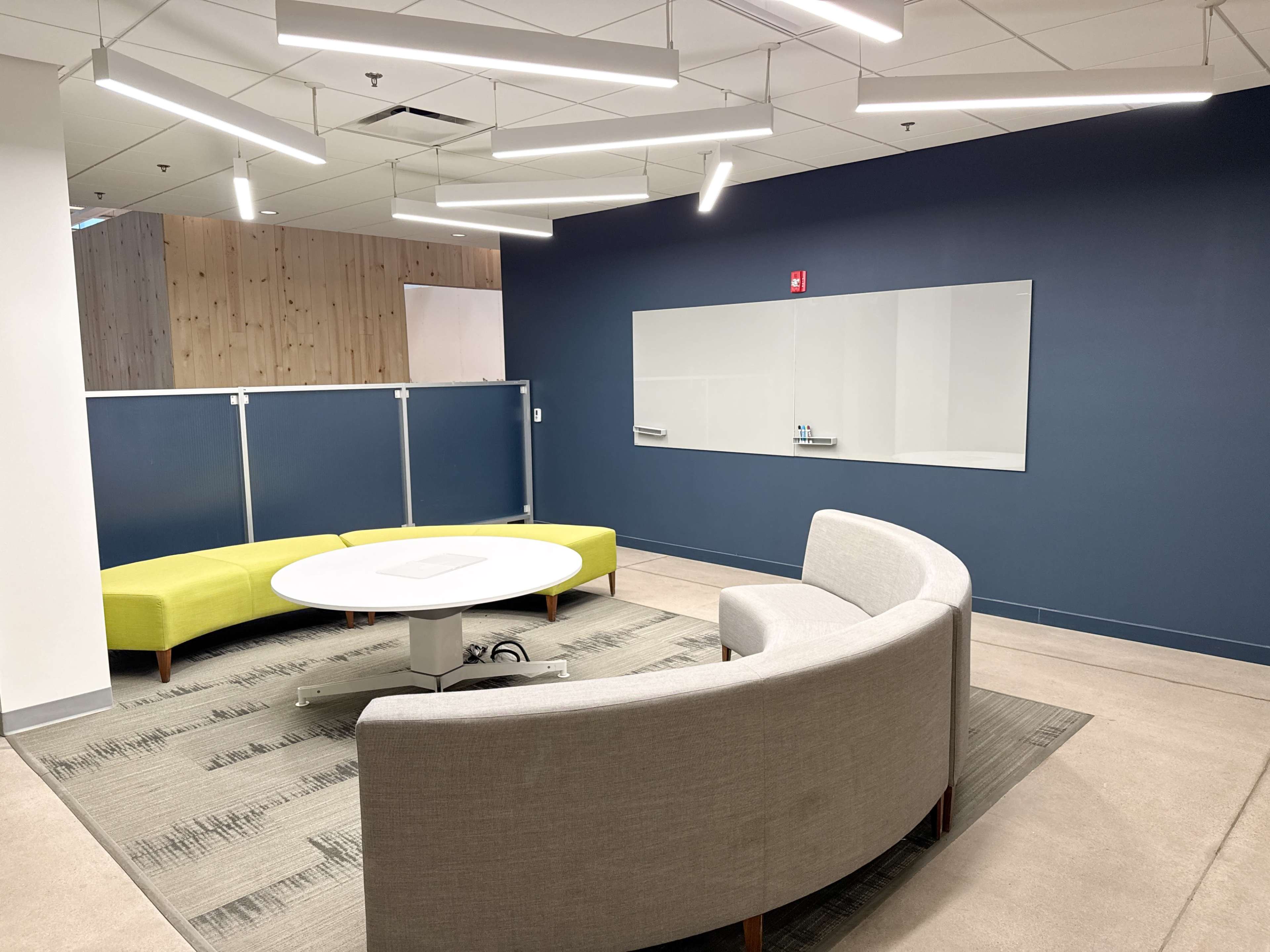 The image shows a modern office space with a circular table surrounded by curved gray seating and a green sofa, against a blue wall with a whiteboard mounted on it.