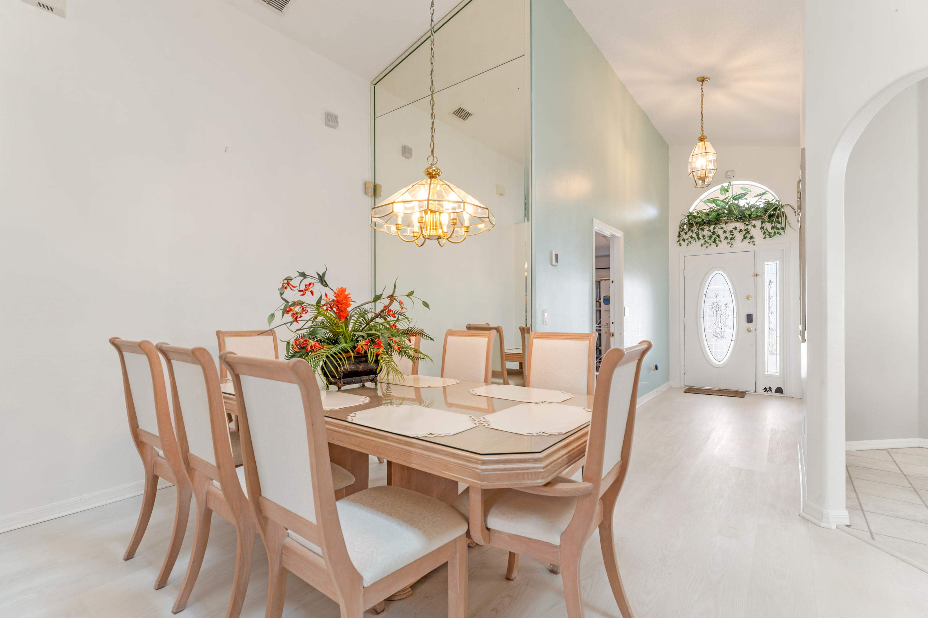 A dining room featuring a wooden table surrounded by eight chairs, with a chandelier overhead and a decorative plant arrangement on the table.