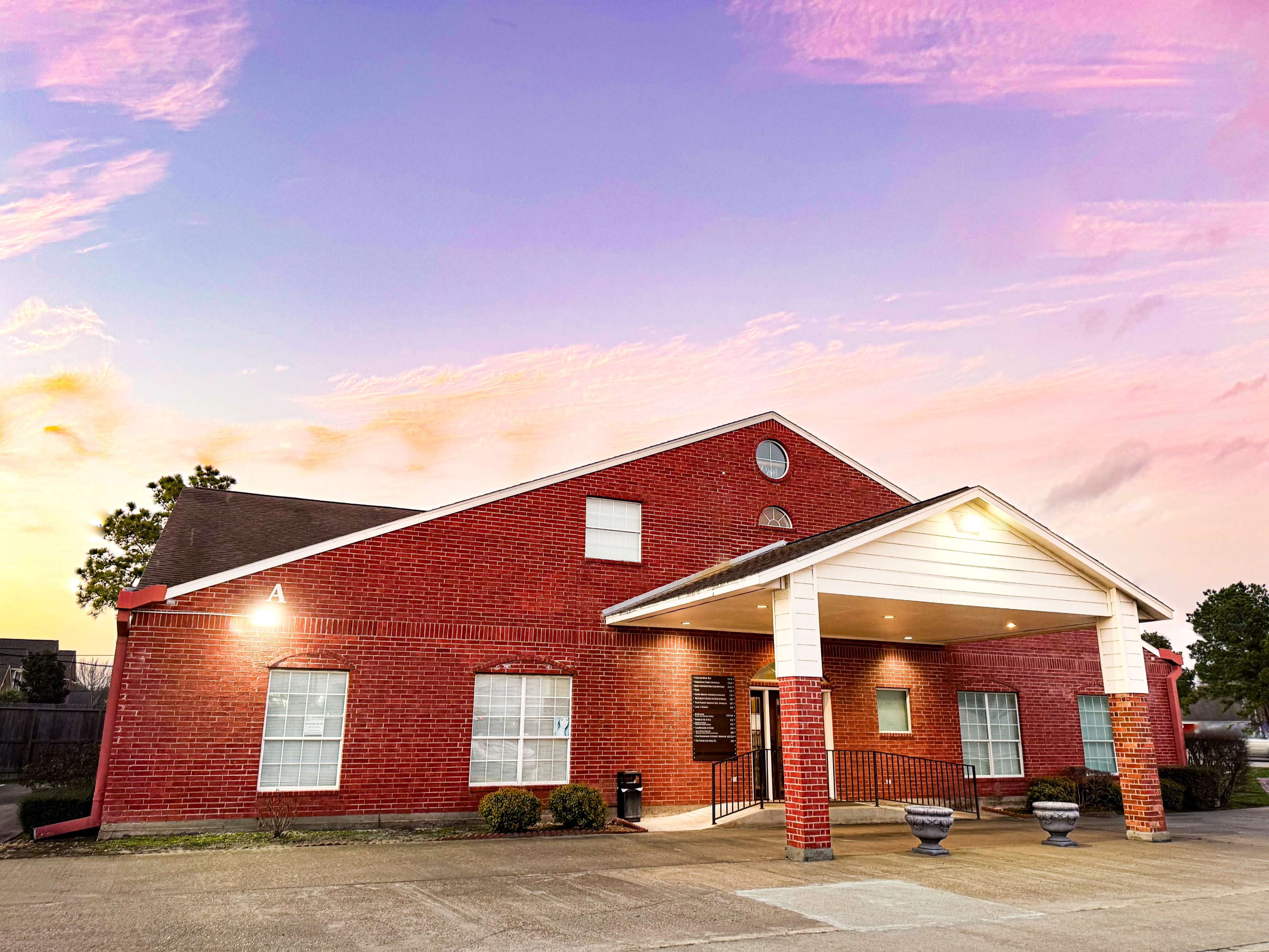 A red brick building with a white entrance and landscaping, set against a colorful sunset sky.