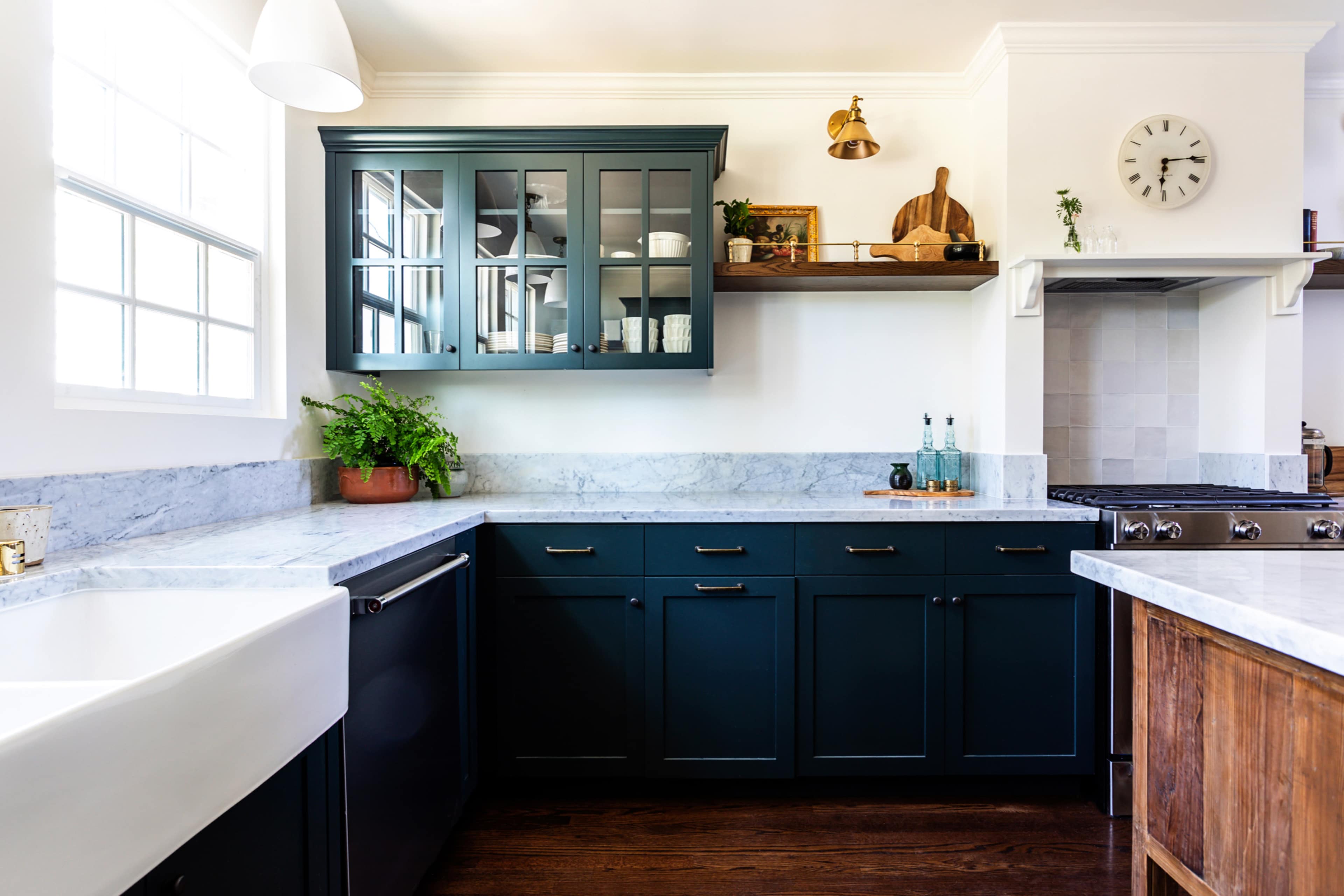A modern kitchen features dark blue cabinetry, a white farmhouse sink, and a marble countertop, complemented by a wooden shelf displaying decorative items.
