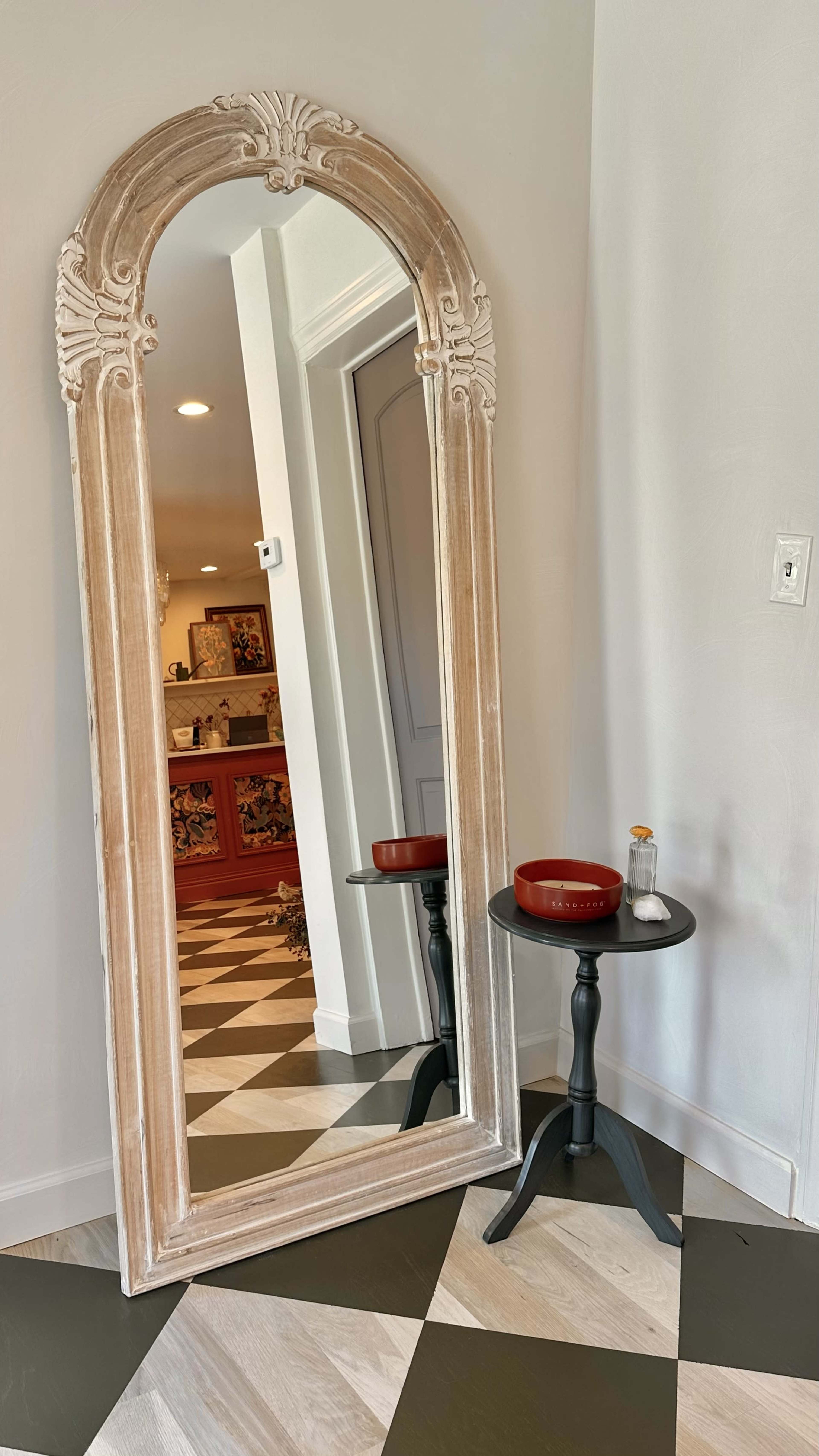 A large, ornate mirror leans against a wall next to a small black table with a round red bowl and a bottle.