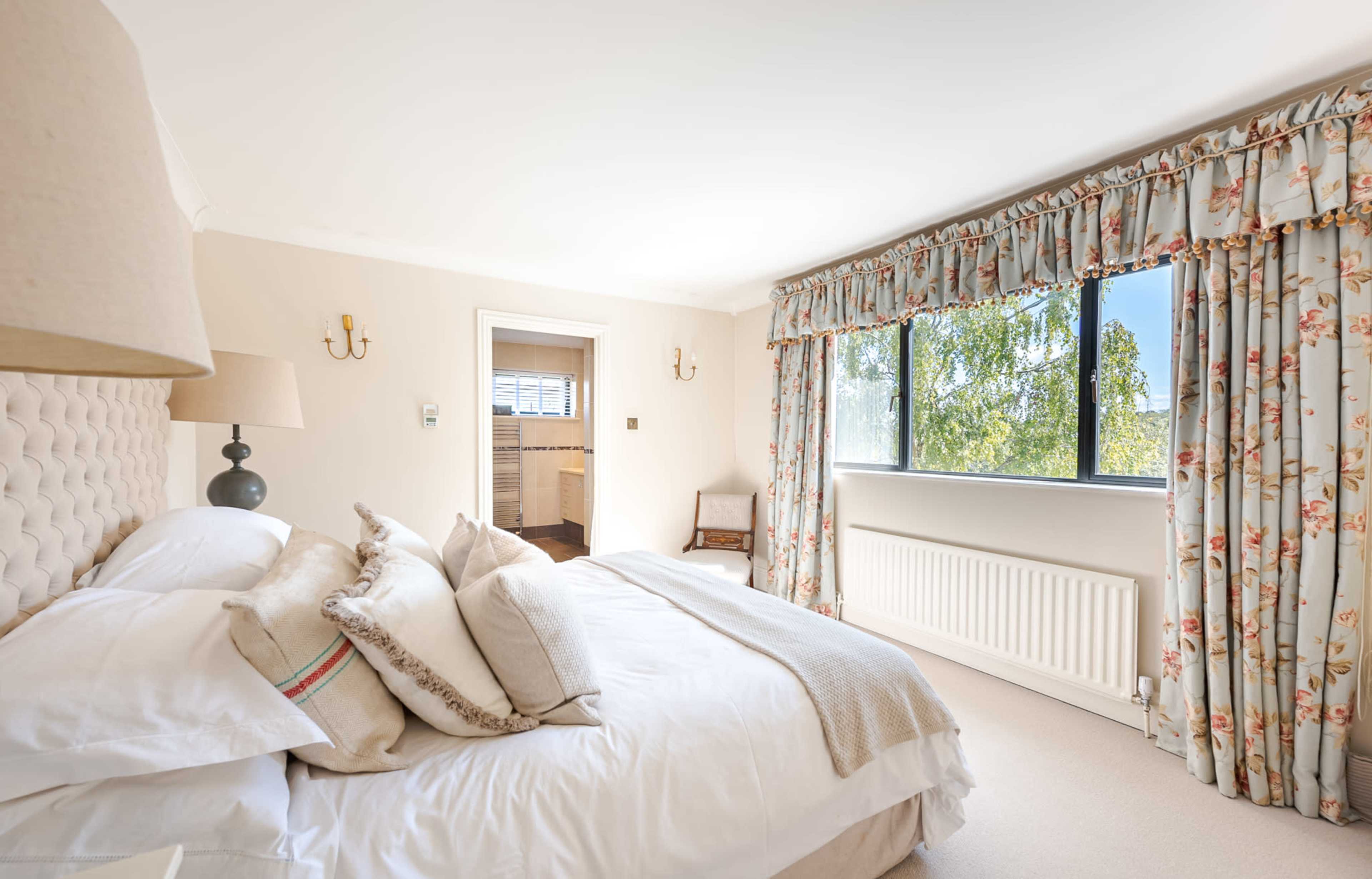 The image shows a softly lit bedroom with a neatly made bed, floral curtains, and a window overlooking a green landscape.