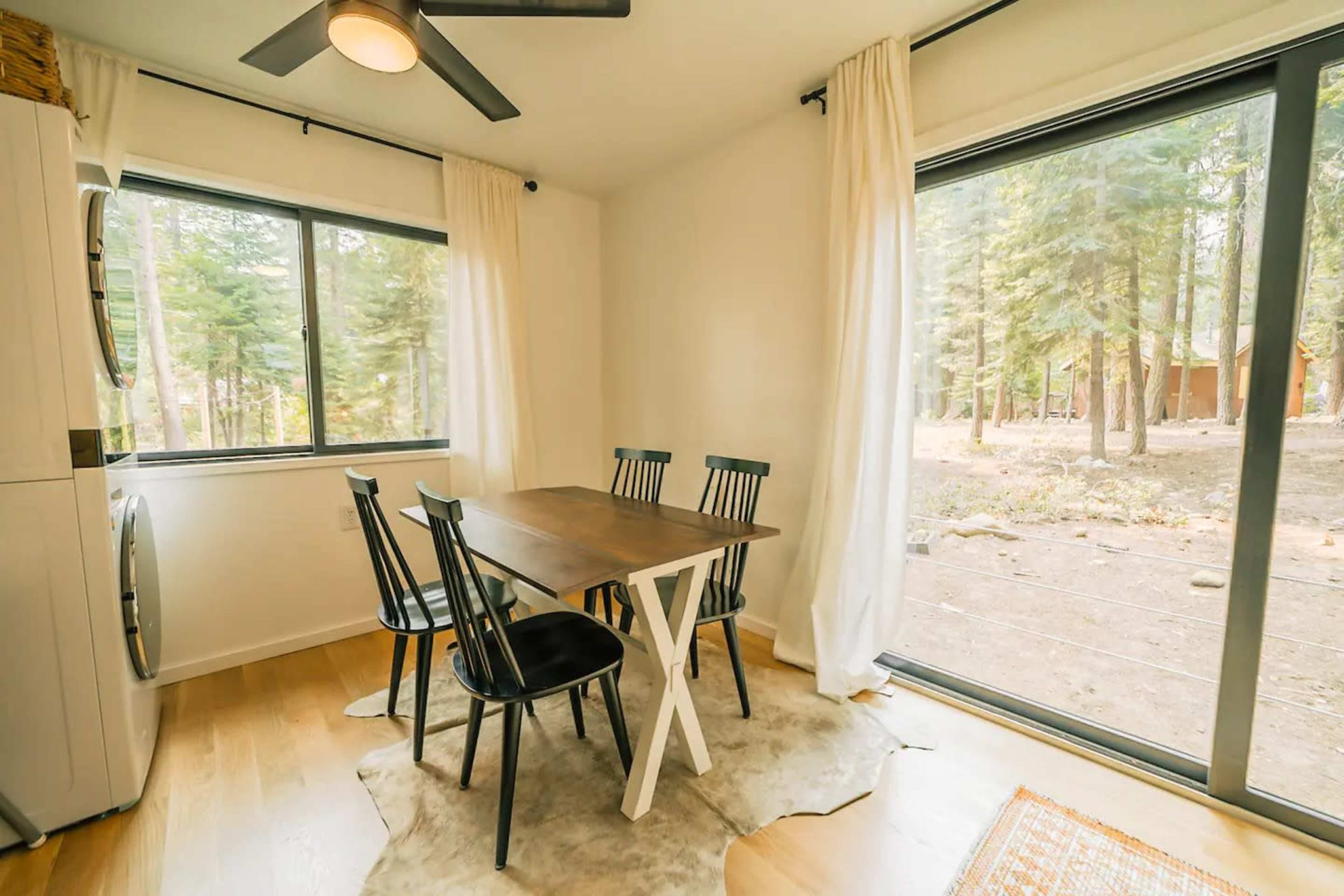 A dining area features a wooden table surrounded by black chairs, with large windows overlooking a wooded landscape.