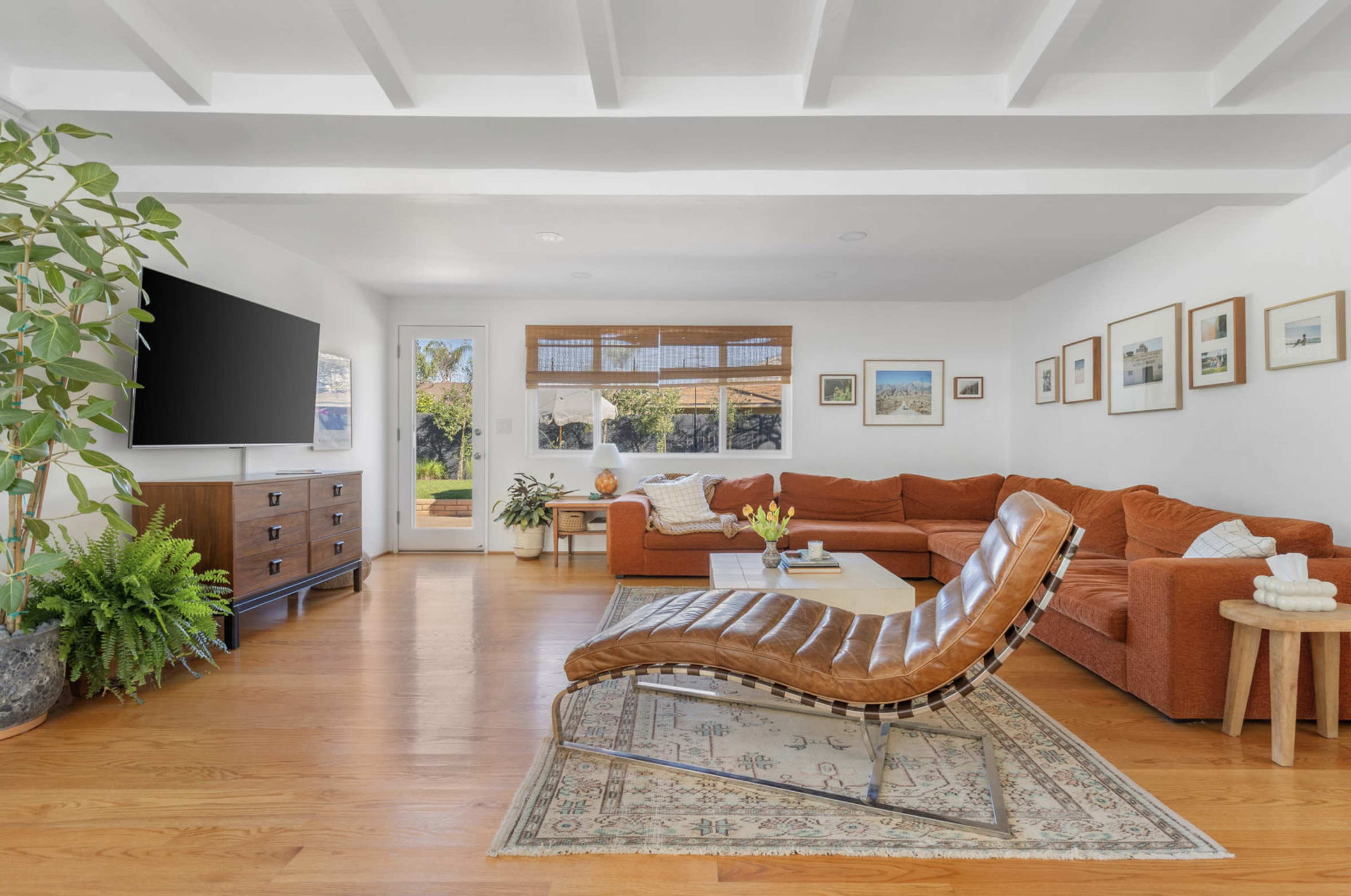 The image shows a bright living room featuring a large orange sectional sofa, a brown leather lounge chair, and a wooden TV console, with large windows allowing natural light and greenery visible outside.