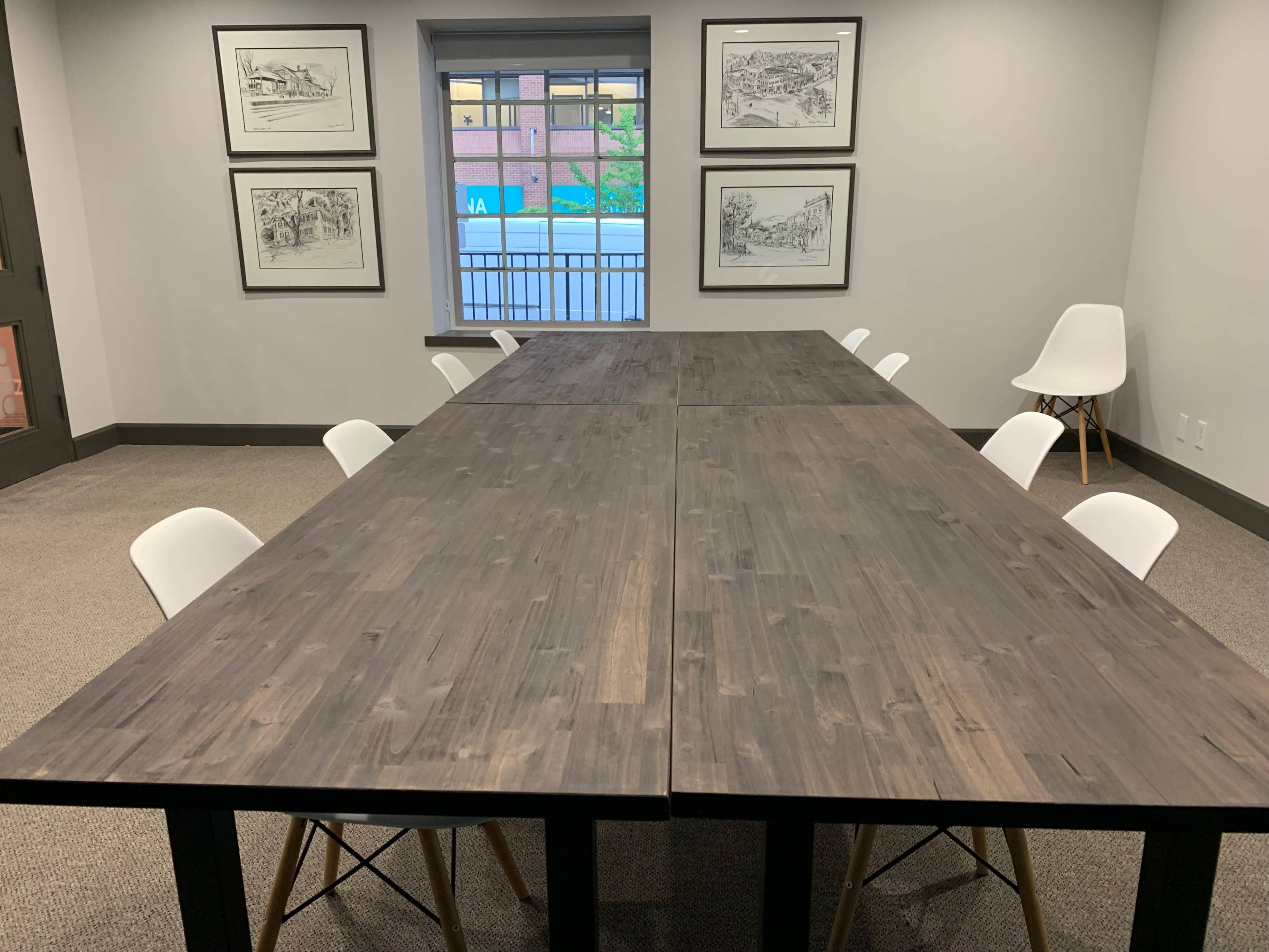 A large, wooden conference table with white chairs is set up in a room featuring framed sketches on the walls and a window in the background.