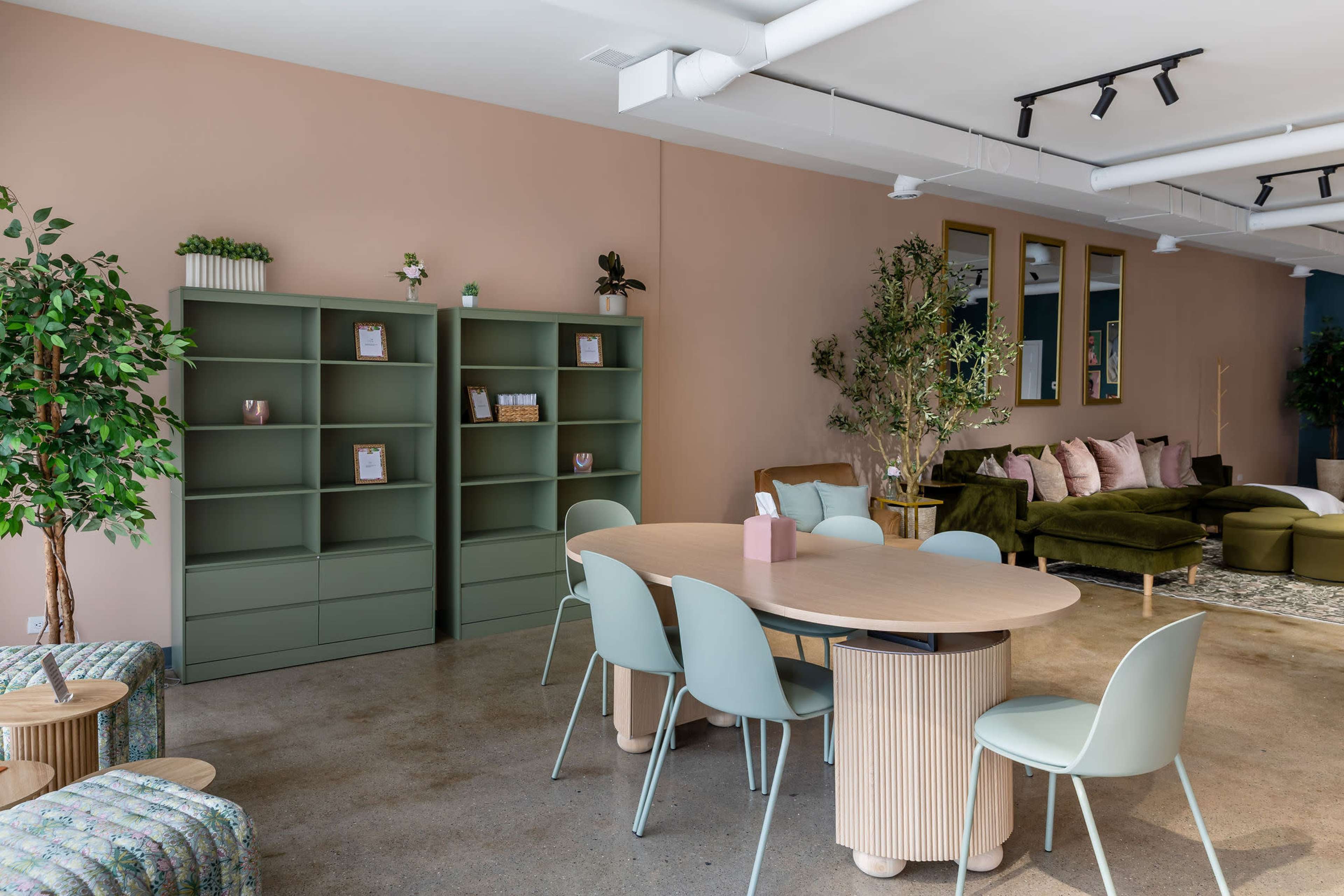 The image shows a modern interior space featuring a round dining table surrounded by pastel-colored chairs, green shelving units, and plush seating arrangements against a pink wall.