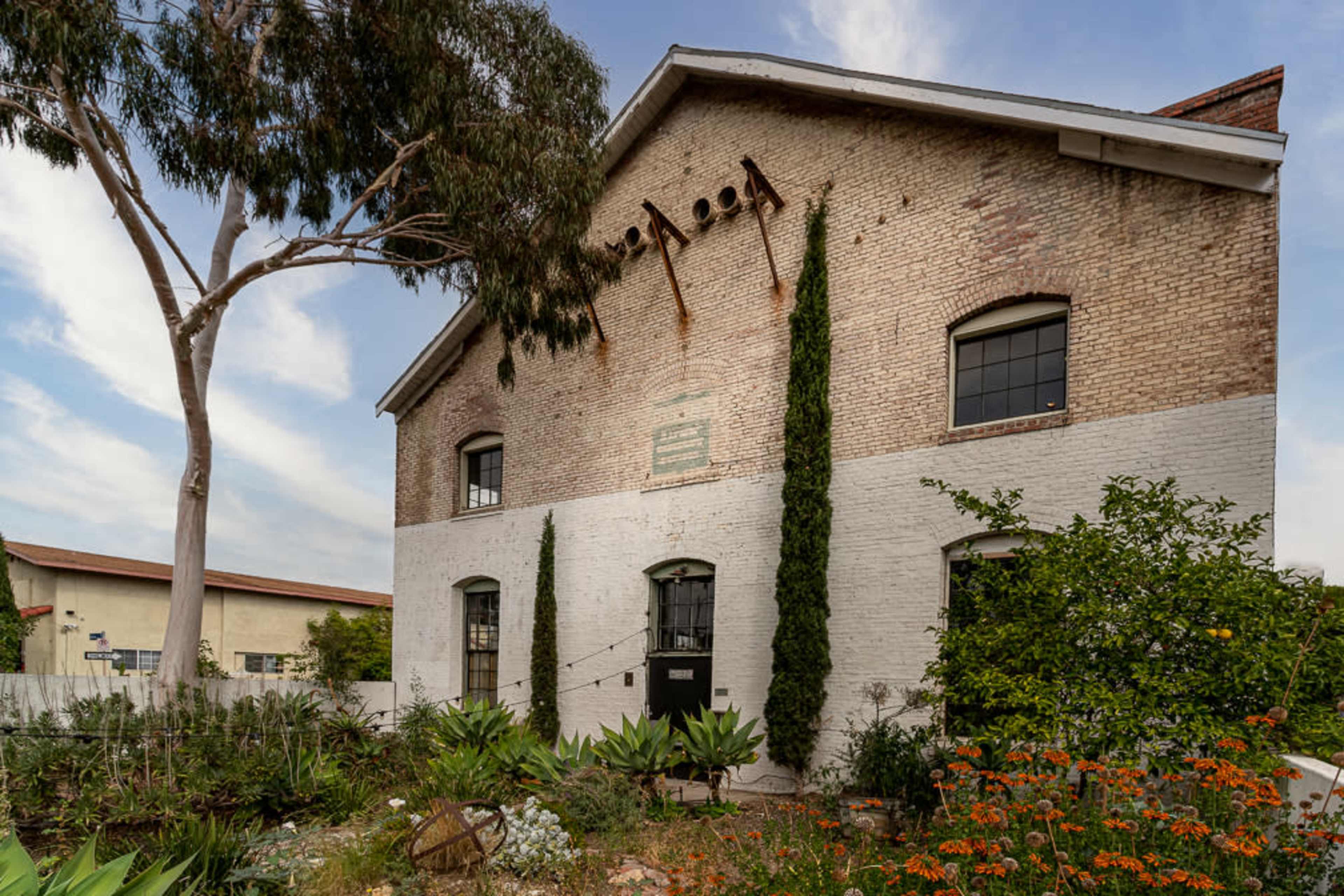 A two-story brick building with tall windows, surrounded by a garden featuring green plants and orange flowers.