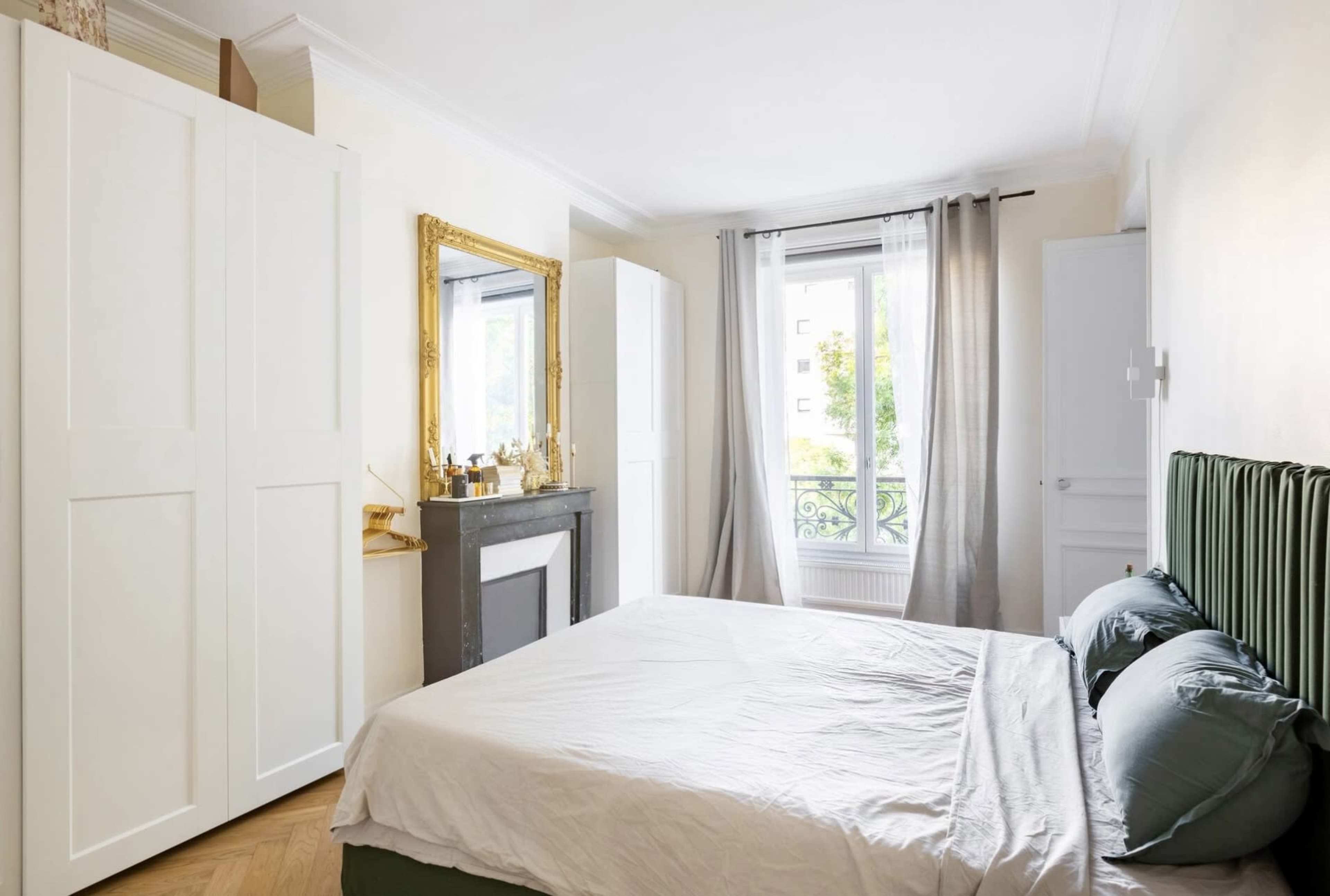The image shows a small, bright bedroom featuring a bed with green and gray linens, a mirror with a gold frame, and a window that offers a view of greenery outside.