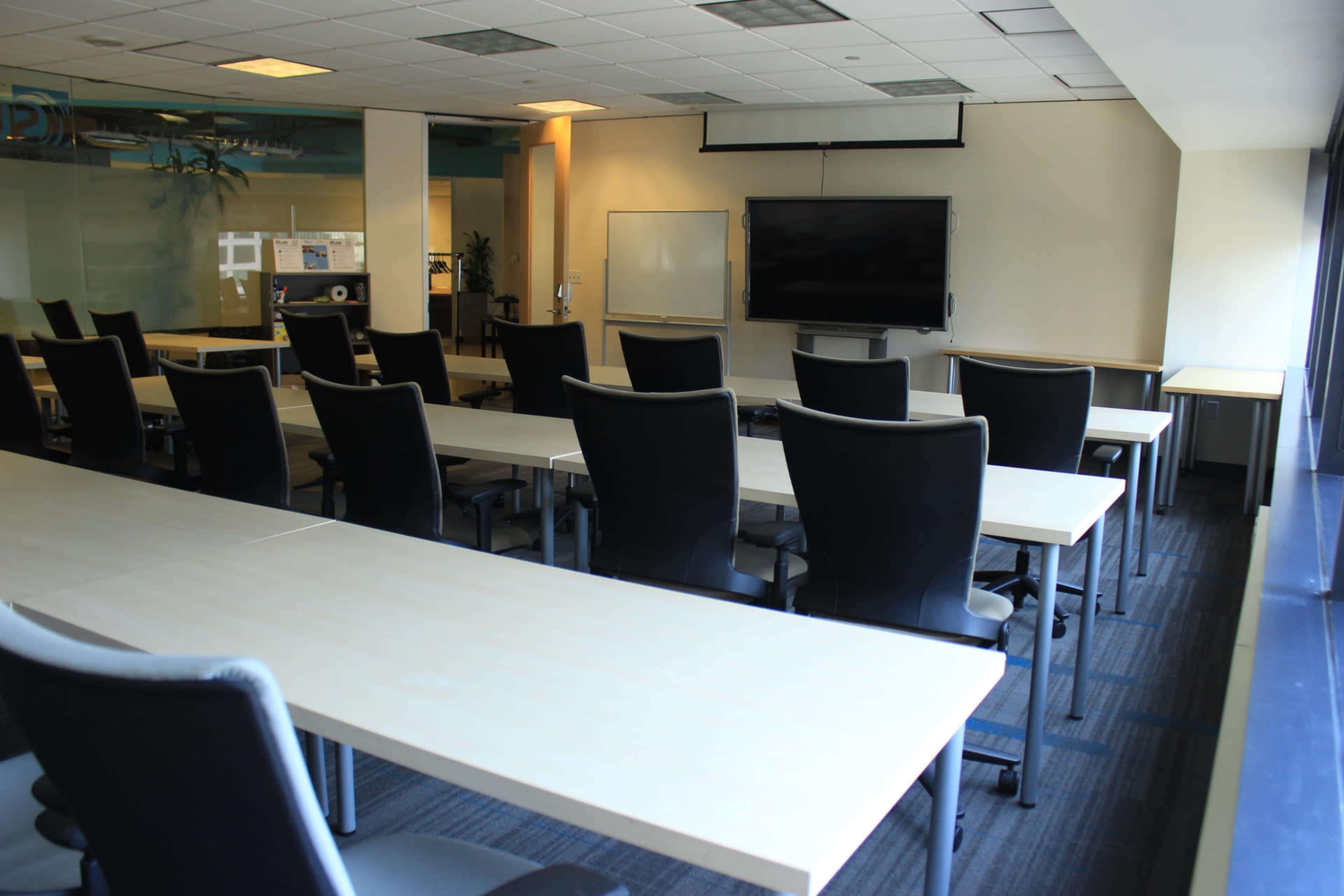 A modern conference room with rows of tables and chairs facing a wall-mounted television screen.