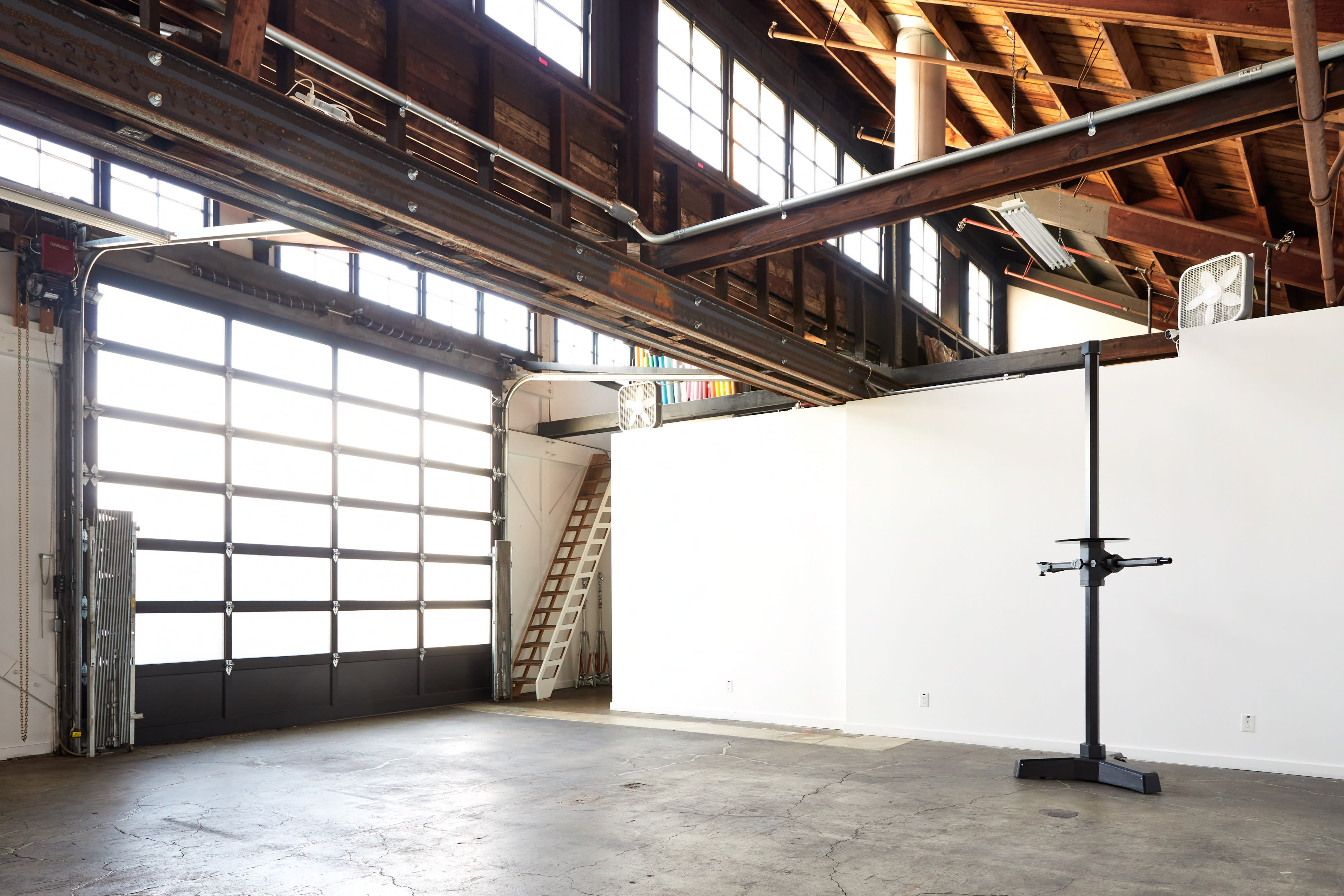 A spacious industrial-style room with large windows, a high ceiling, and a loading dock door, featuring a blank white wall and a mounted display stand.