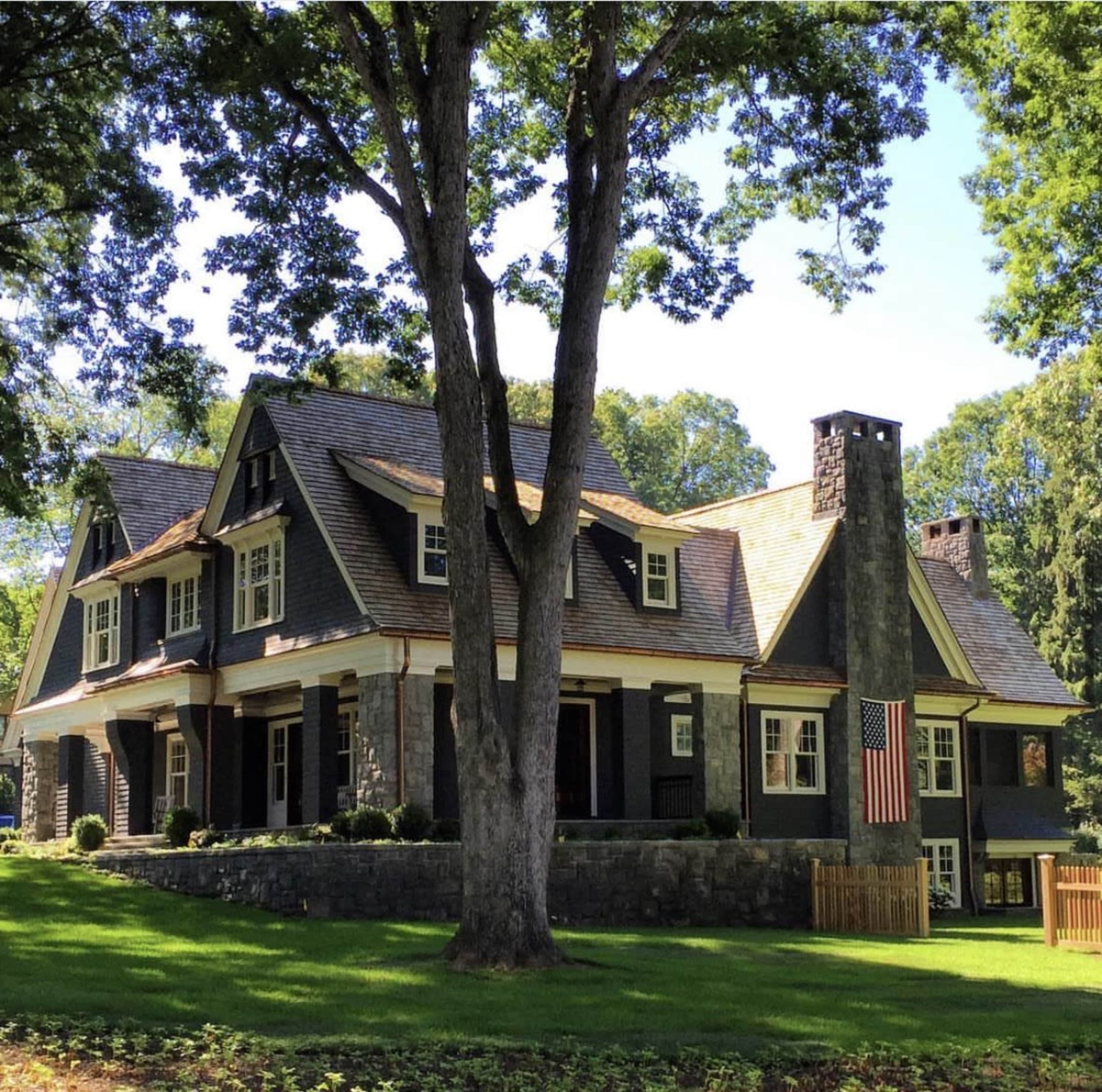 The image shows a large, two-story house with a stone exterior, an American flag on the porch, and surrounded by trees and a well-maintained lawn.