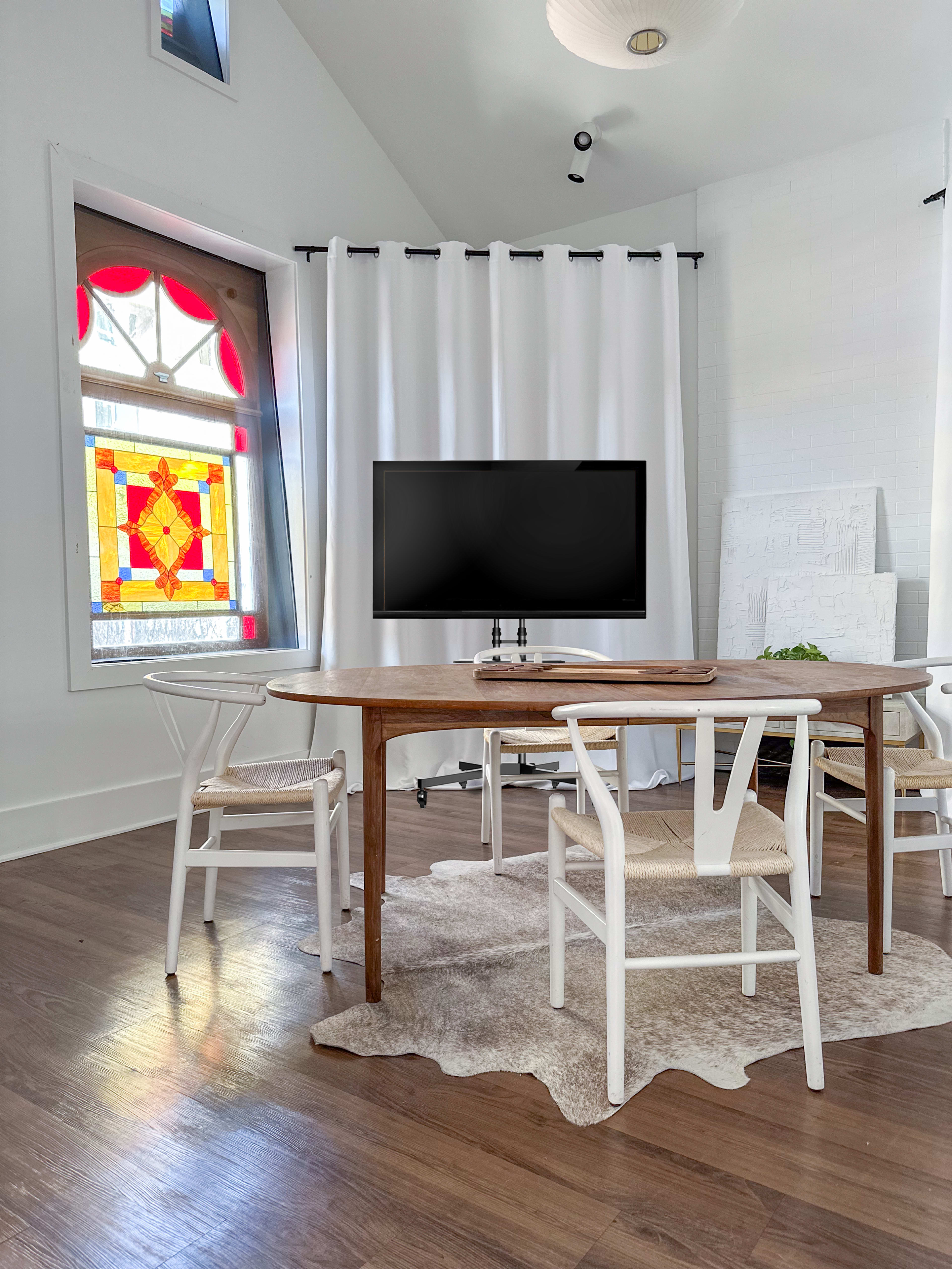 A wooden dining table surrounded by white chairs sits in a well-lit room with a large window featuring a colorful stained glass design and a television mounted on the wall.