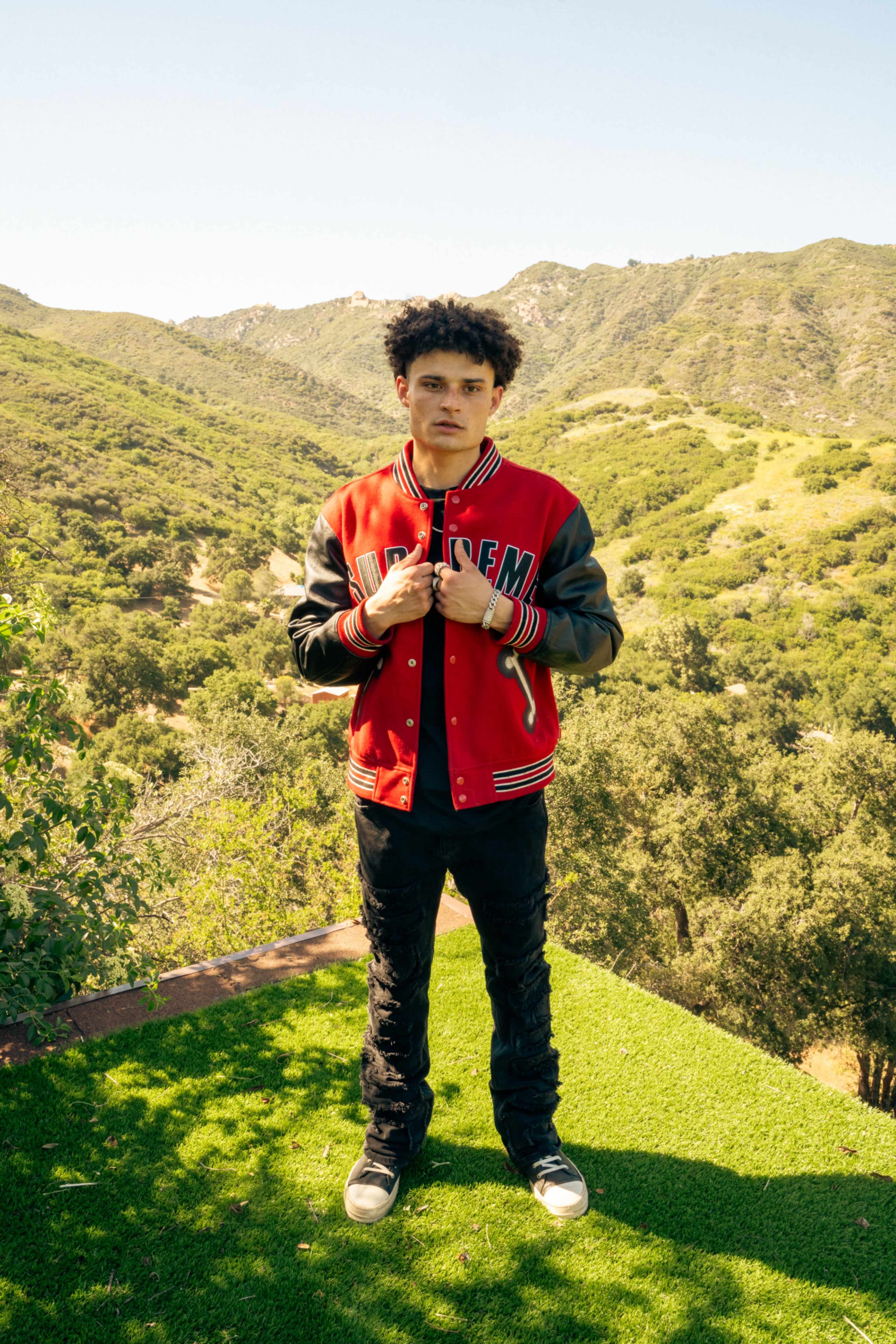 A person stands on a grassy area with hills covered in green vegetation in the background.