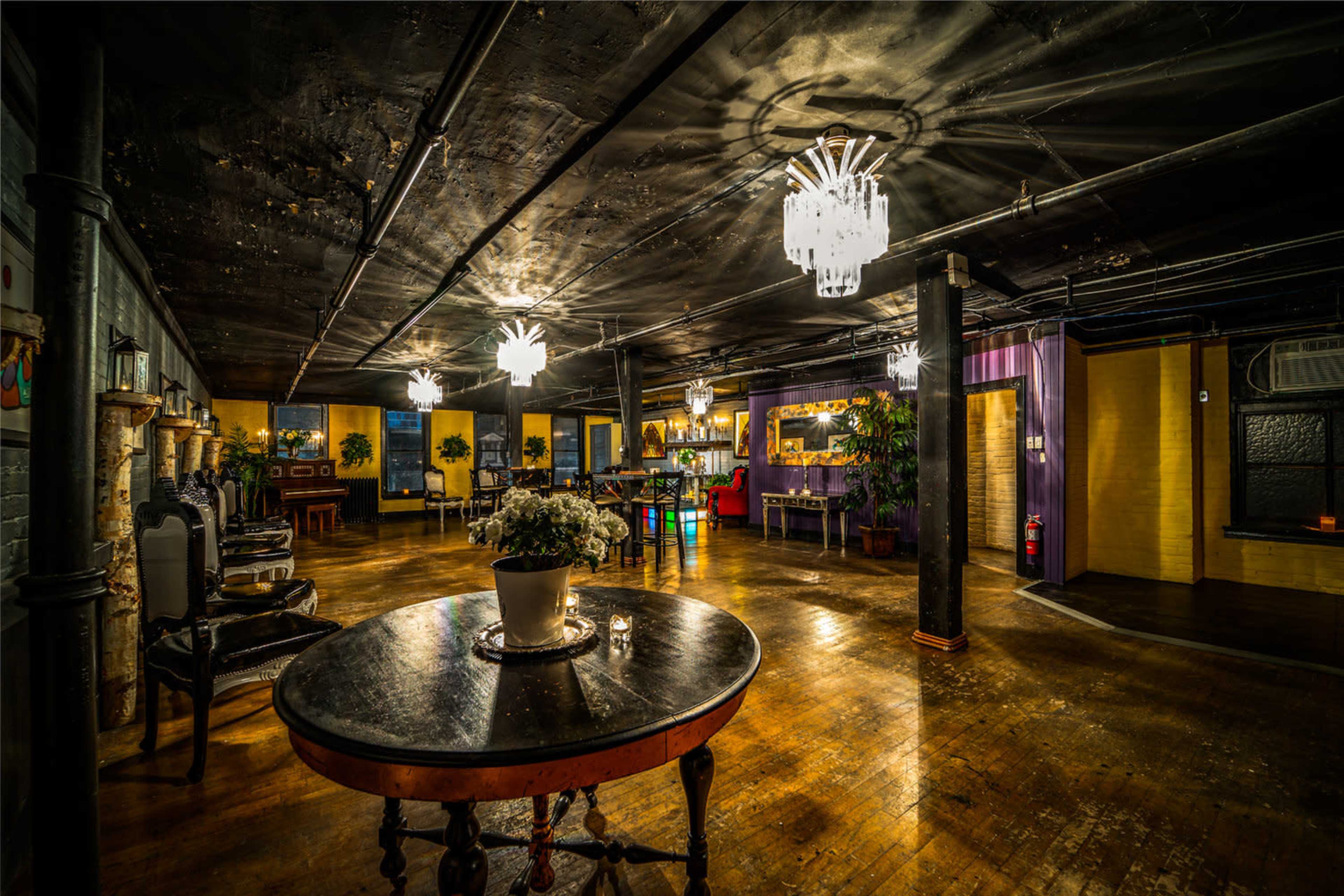 A dimly lit, spacious interior featuring a wooden floor, elegant chandeliers, and various seating arrangements.
