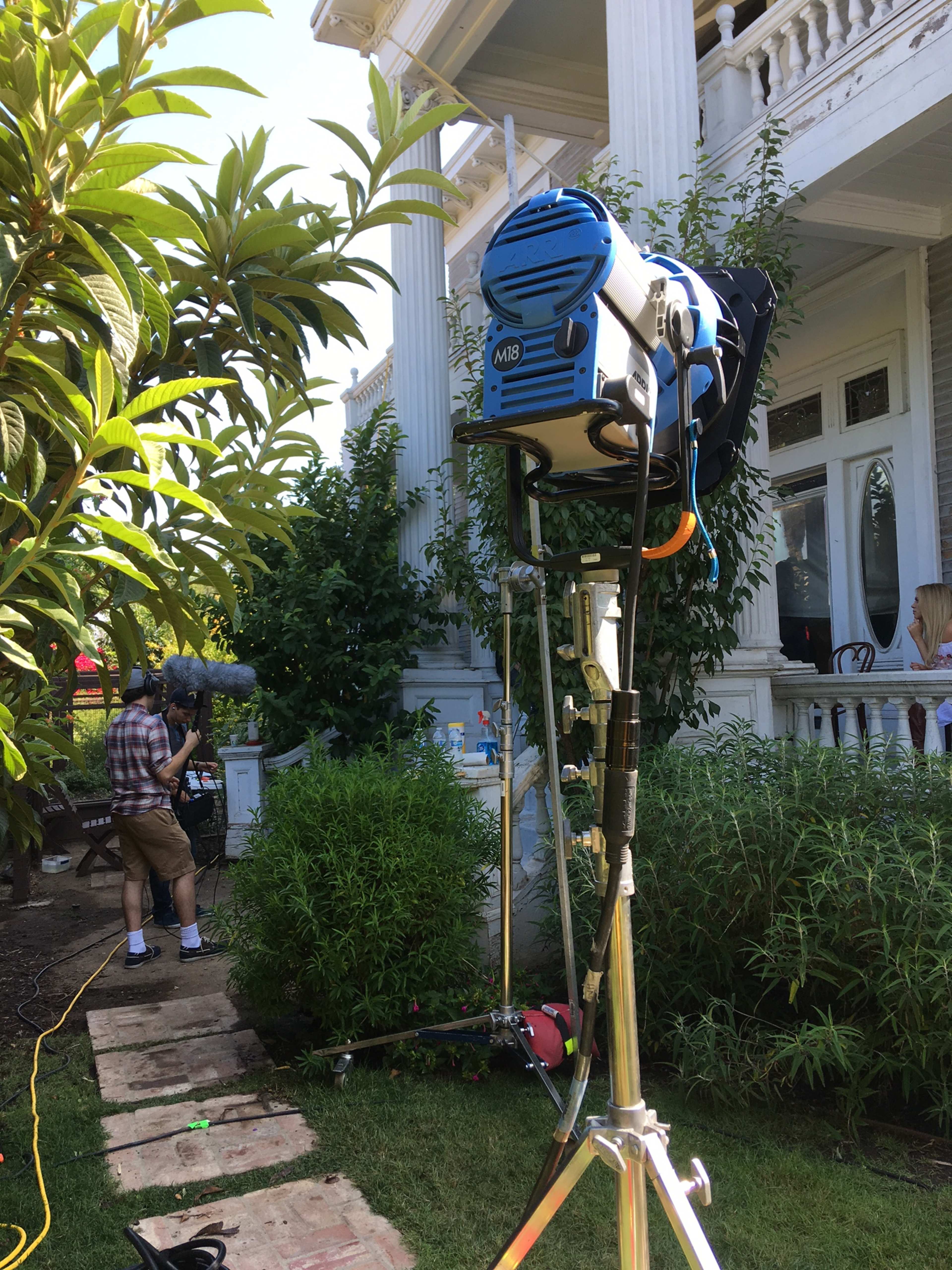 A film crew is set up outside a large, historic house, with a light mounted on a stand and a person operating sound equipment nearby.